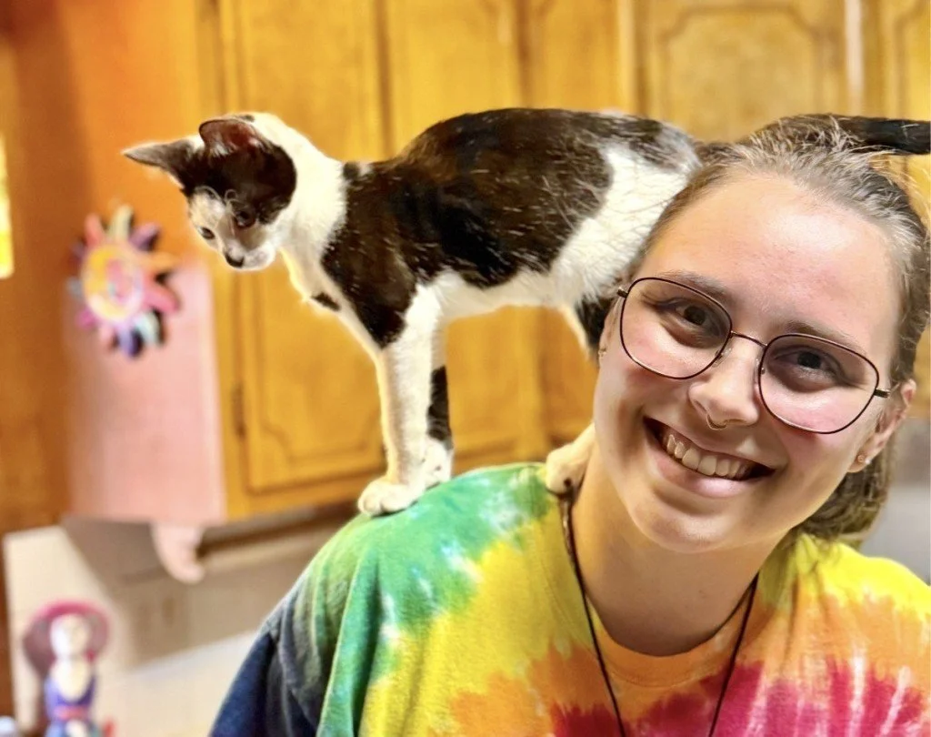 A young woman with glasses and a rainbow-colored tie-dye shirt smiling, with a black and white kitten standing on her shoulder. The background shows a wooden kitchen cabinet.