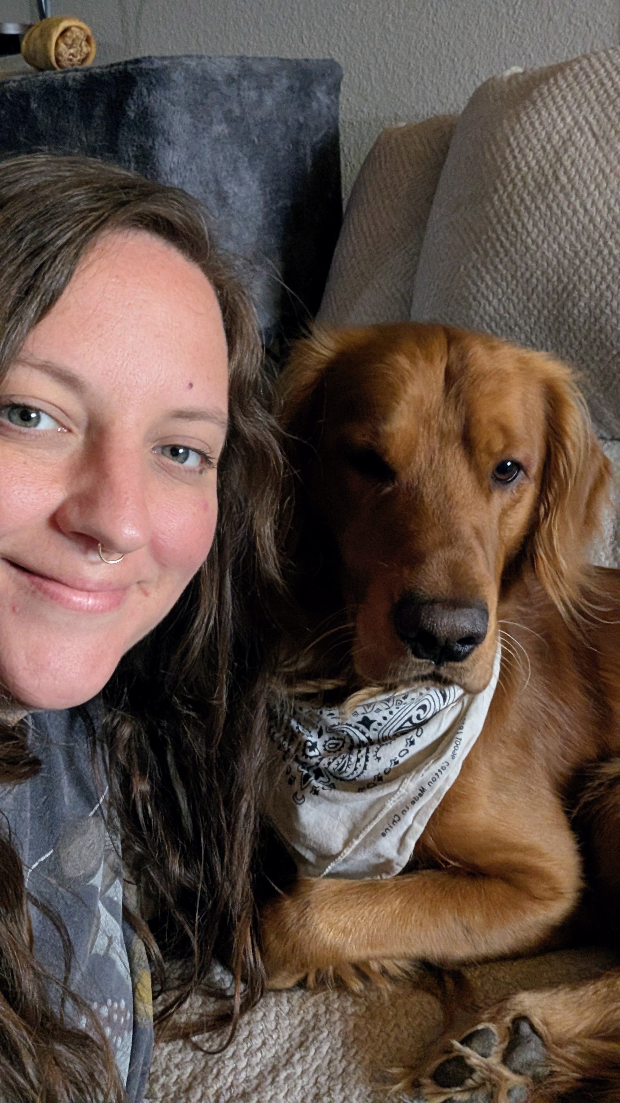 A woman with long, curly hair and a septum piercing smiling next to a golden retriever dog lying on a carpeted floor, wearing a bandana around its neck.