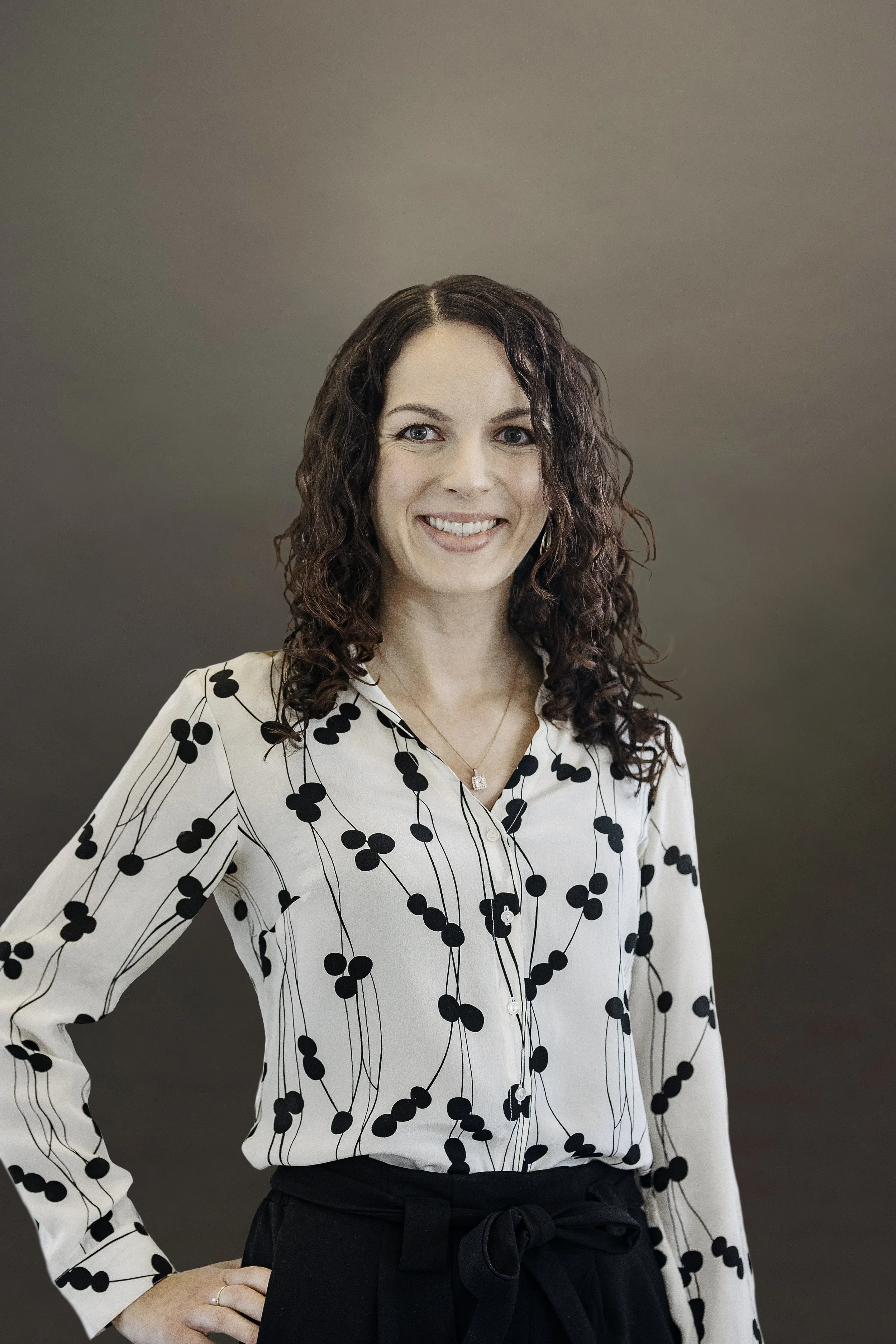 A woman with curly dark hair wearing a white blouse with black dot pattern and black high-waisted pants, standing with one hand on her hip and smiling.