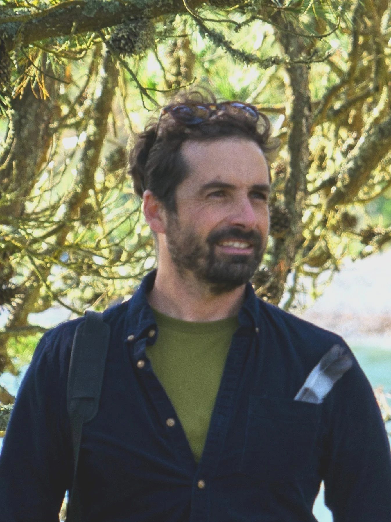 A man with a beard wearing a dark shirt and green undershirt stands in a wooded area with sunlight filtering through the trees.