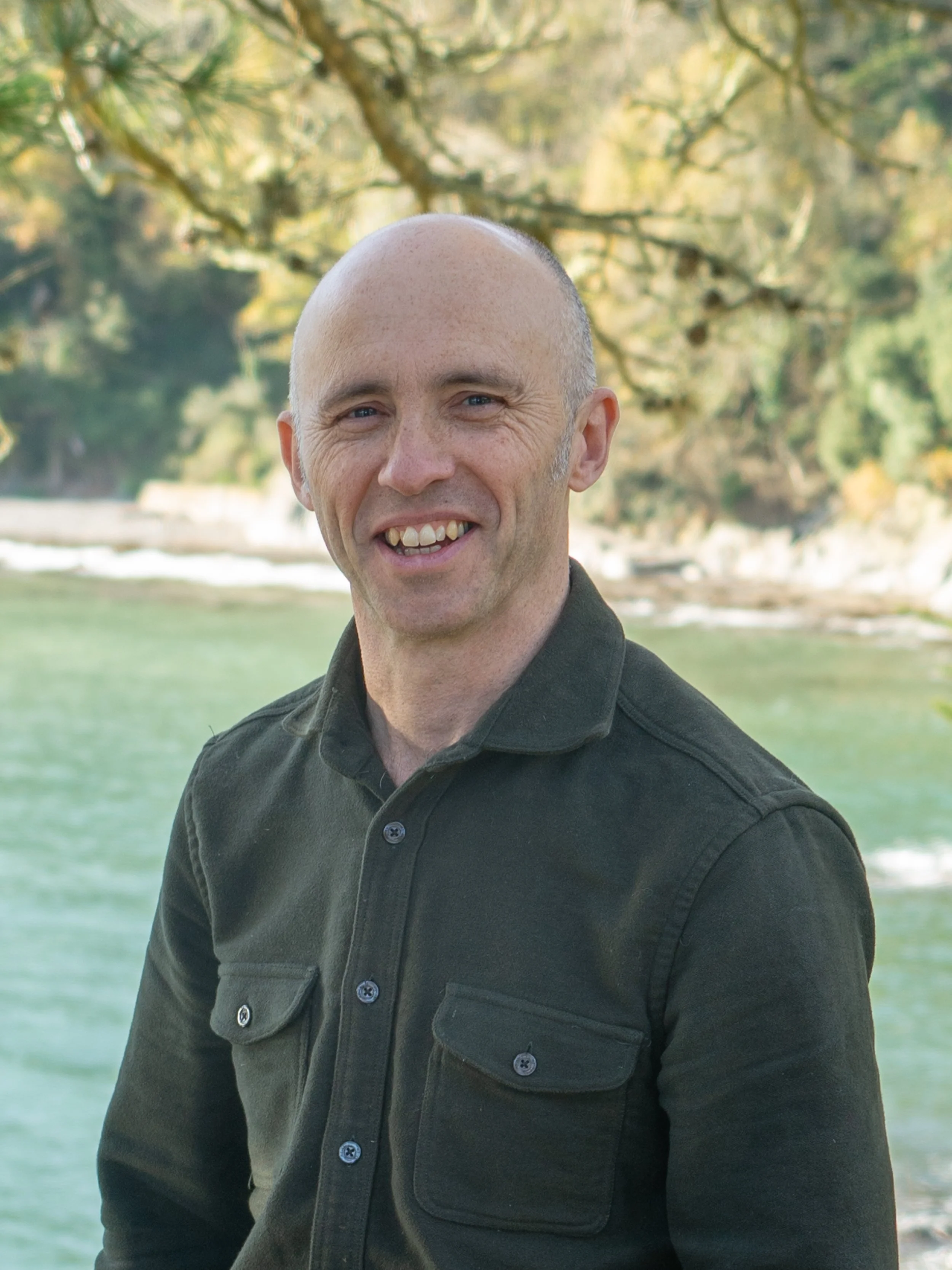 Man in green jacket smiling outdoors near water with trees in the background.