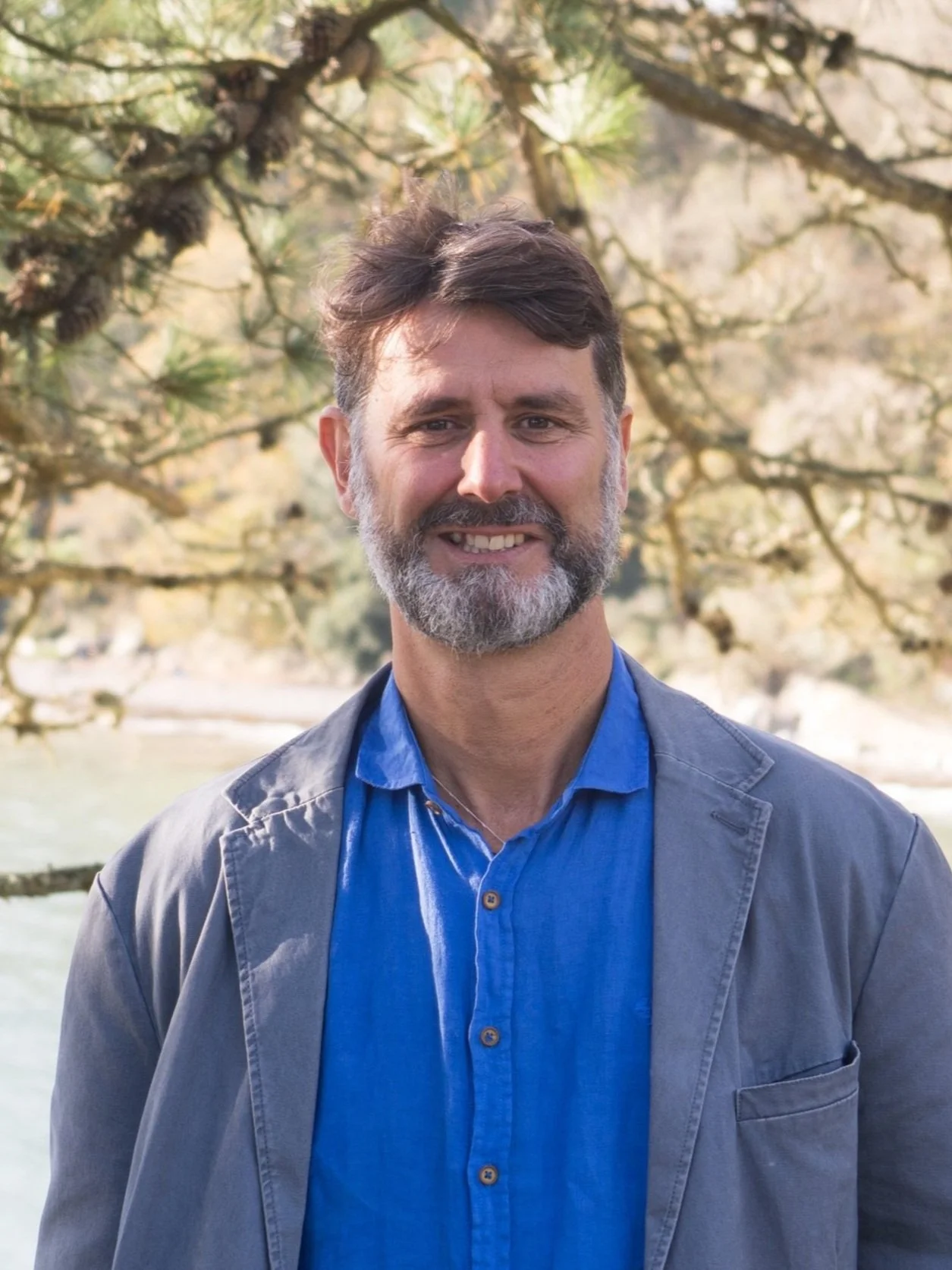 A man with a beard and mustache smiling, wearing a blue shirt and gray jacket, standing outdoors in front of a tree.