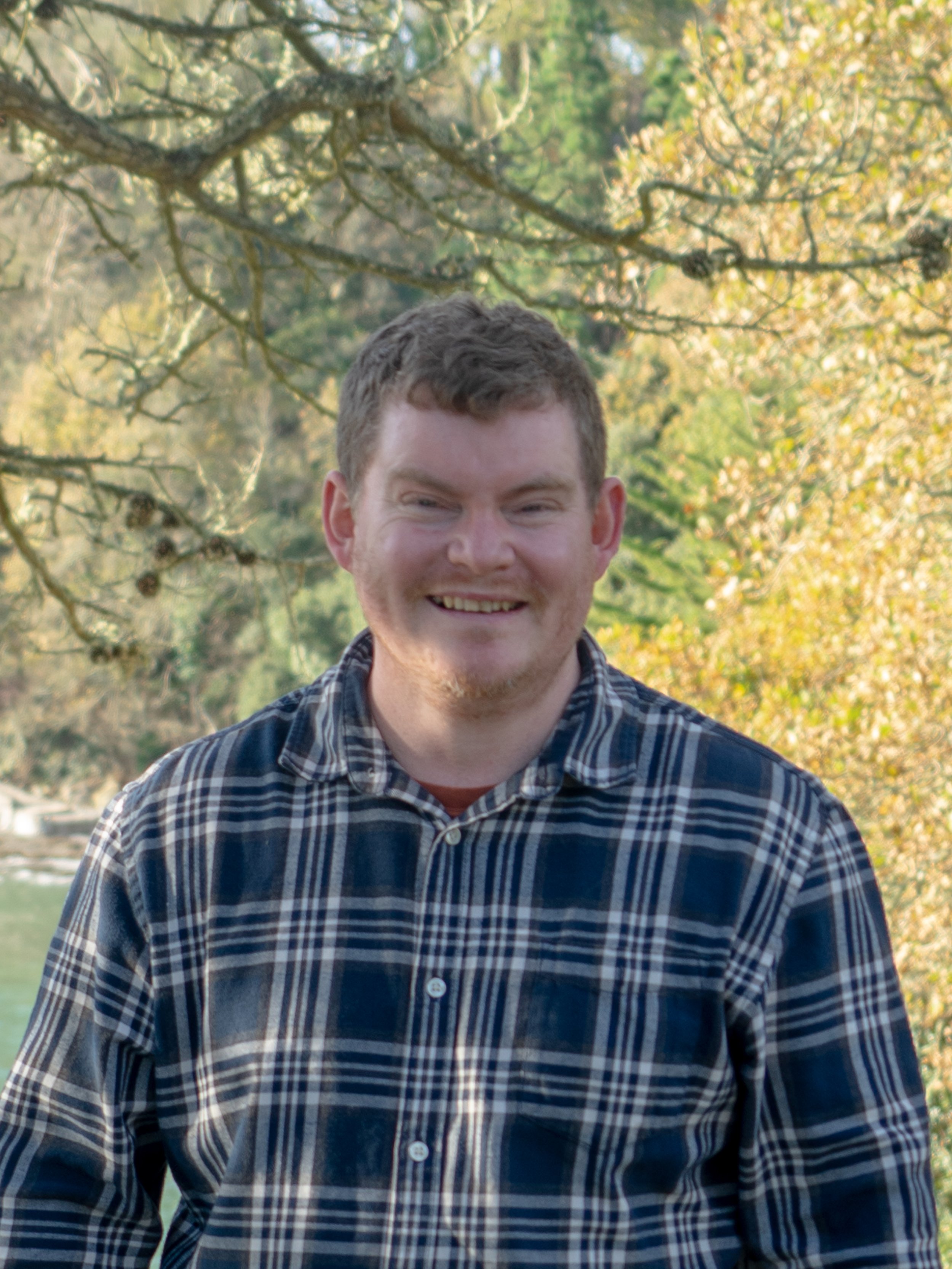Man in a plaid shirt standing outdoors with trees in the background