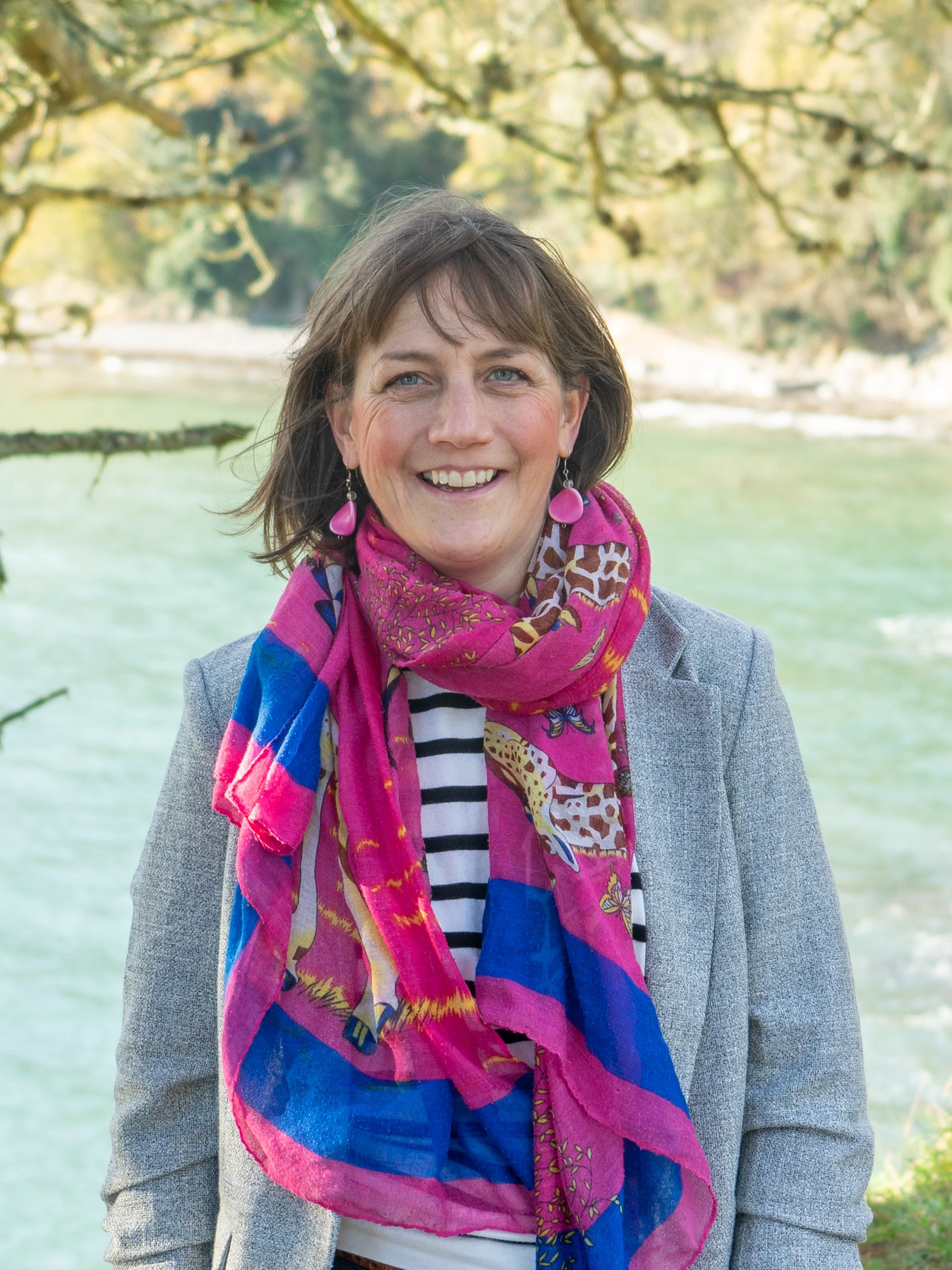 Smiling person with short hair wearing a colorful scarf and blazer, standing outdoors near a body of water and trees.
