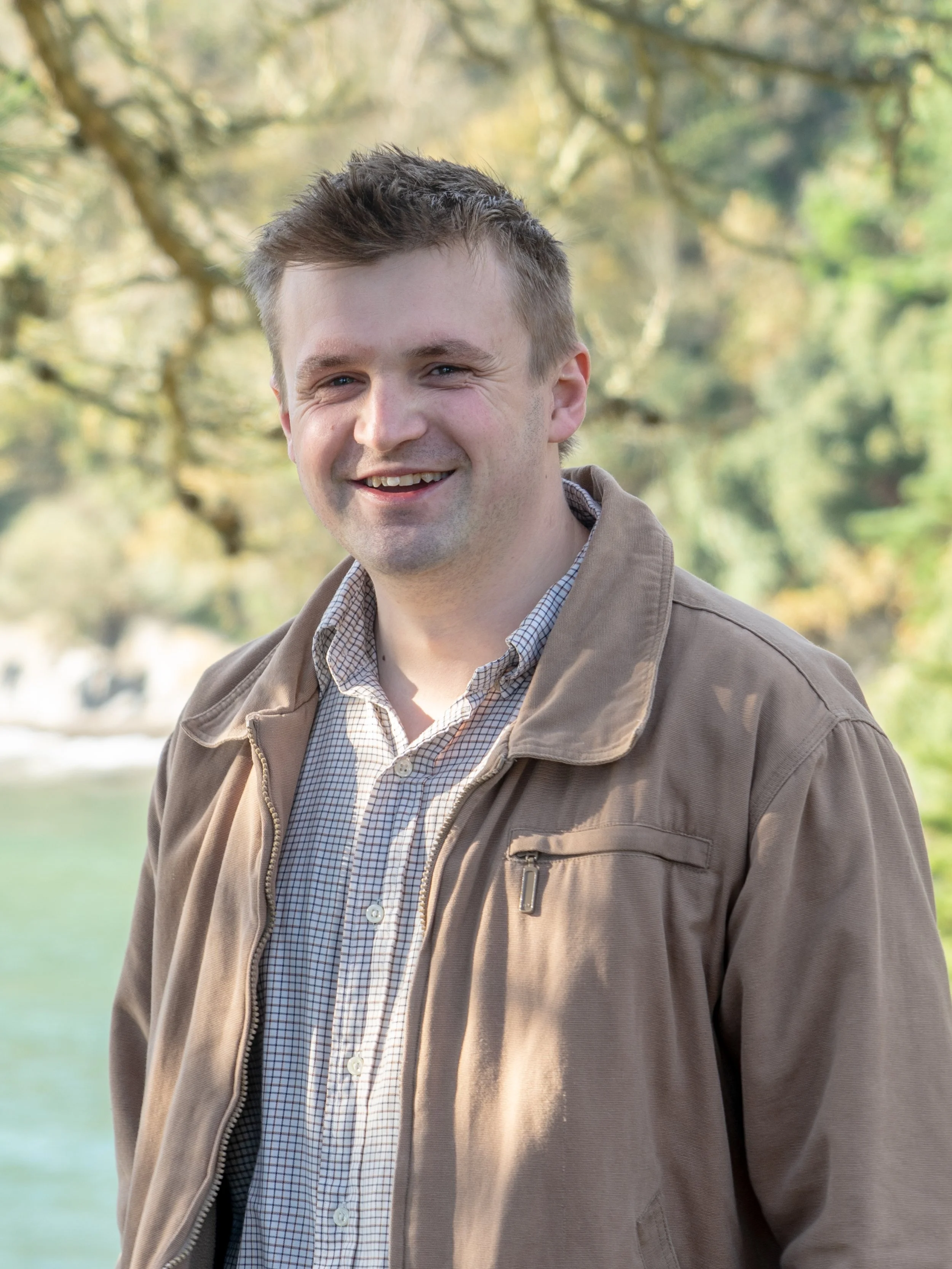 Person wearing a brown jacket and checkered shirt, standing outdoors in a nature setting with trees and water in the background.