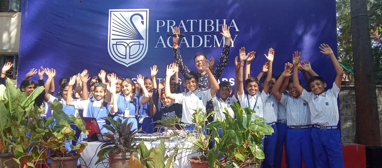 Group of school children and a teacher standing outdoors, smiling, and raising their hands in front of a blue banner for Pratibha Academy.