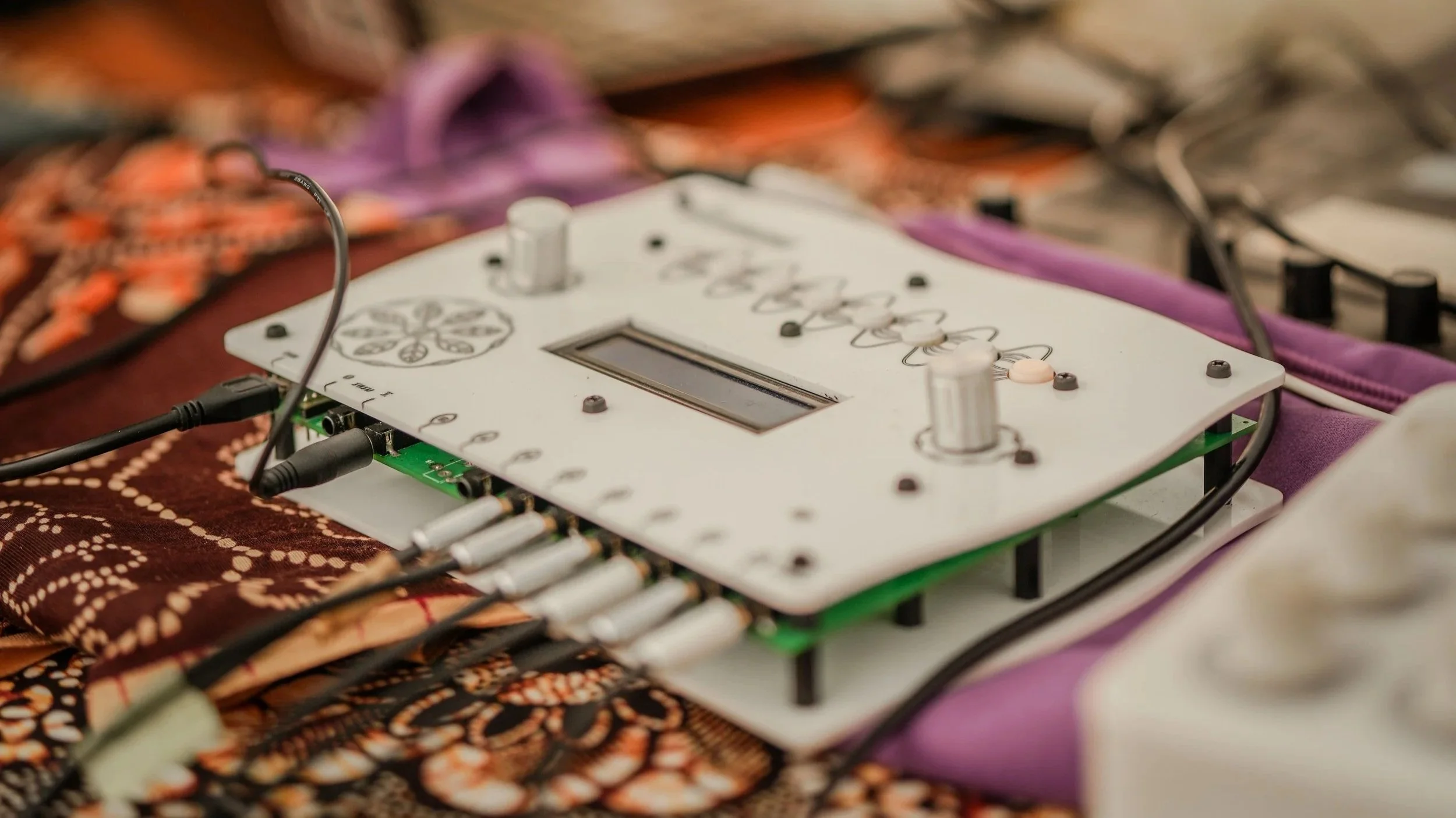 Electronic musical instrument or synthesizer with multiple knobs and a small display screen, connected to various cables, placed on patterned fabric.