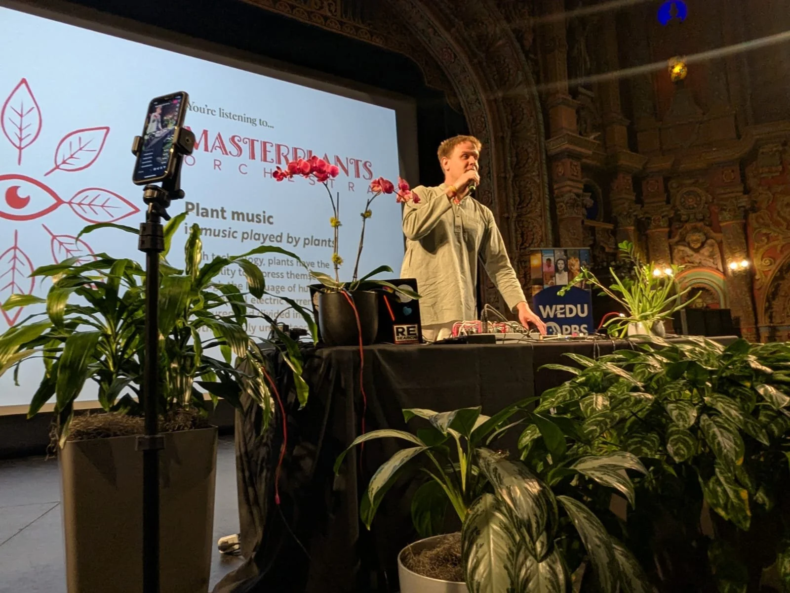 A man in a light shirt sits on the floor in a dimly lit space with candles and plants, operating electronic music equipment.