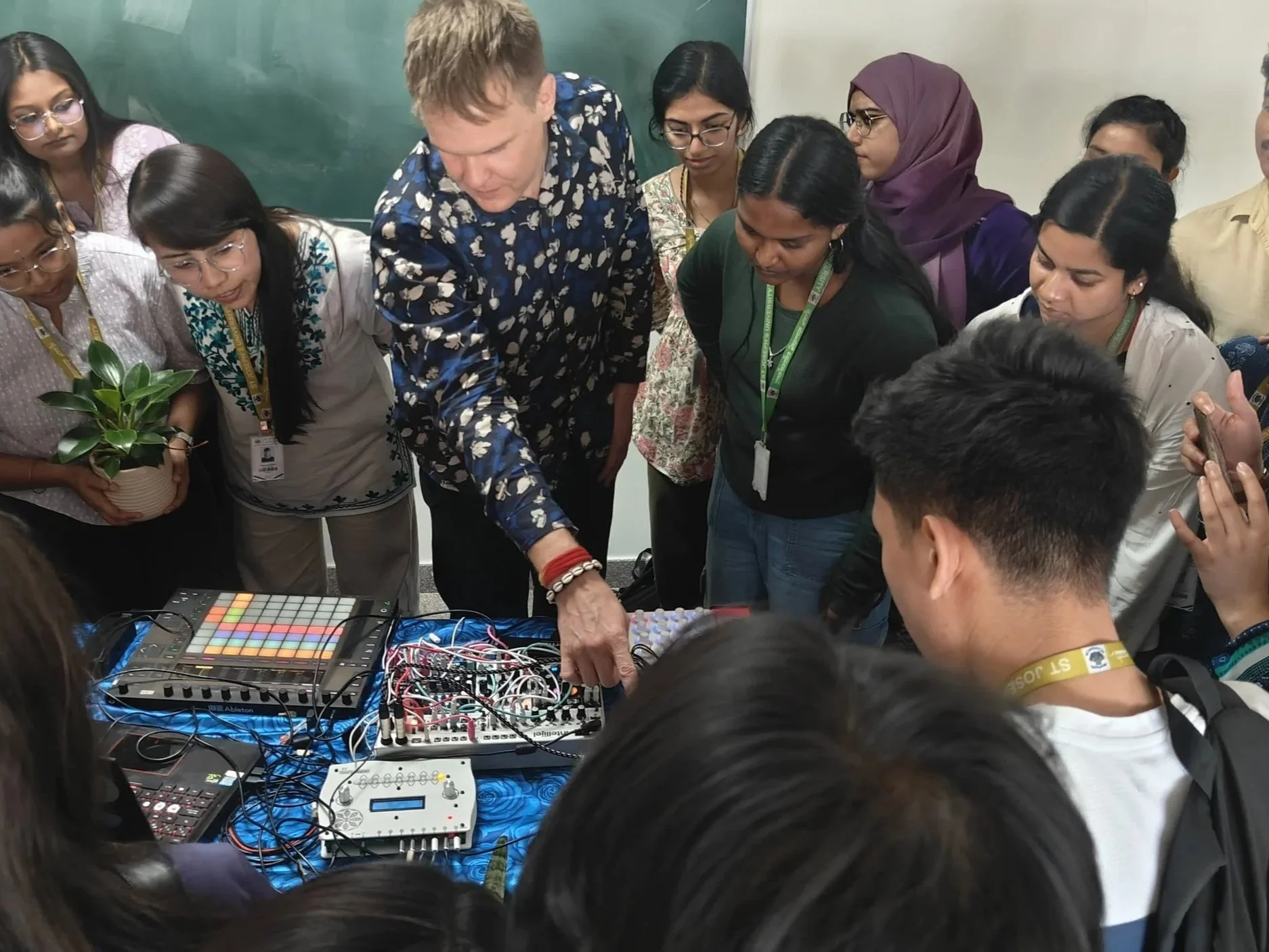 A group of people gathered around a table with electronic music equipment, including a drum pad, mixer, and various cables, as a person demonstrates how to use the equipment.