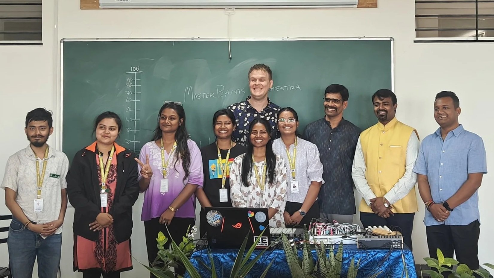 A group of ten people standing in front of a classroom blackboard, smiling for the photo. The blackboard has some writing and a height chart. There are plants and equipment on a table in front of the group.