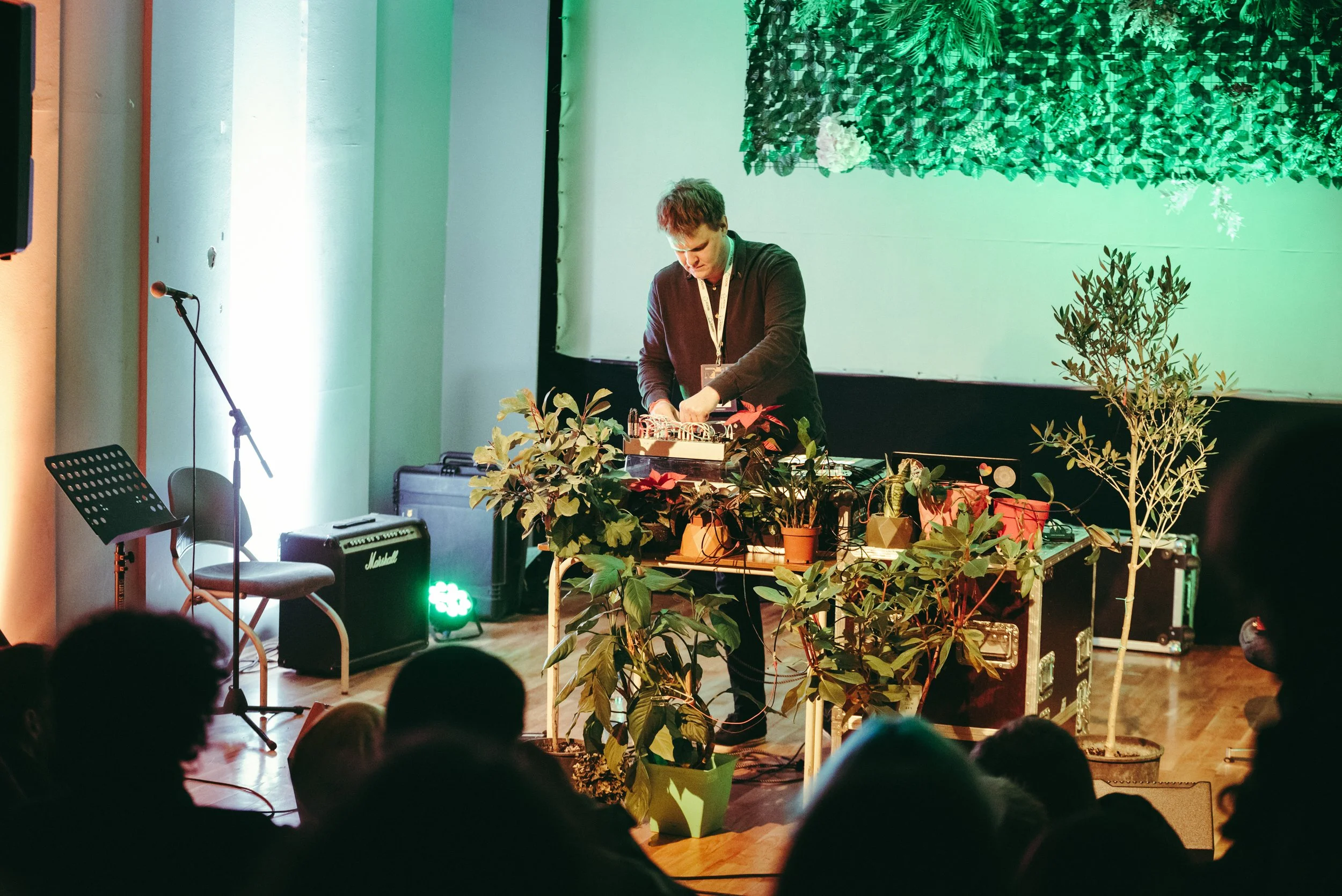 A DJ performing on stage with plants around him, and an audience watching in front.
