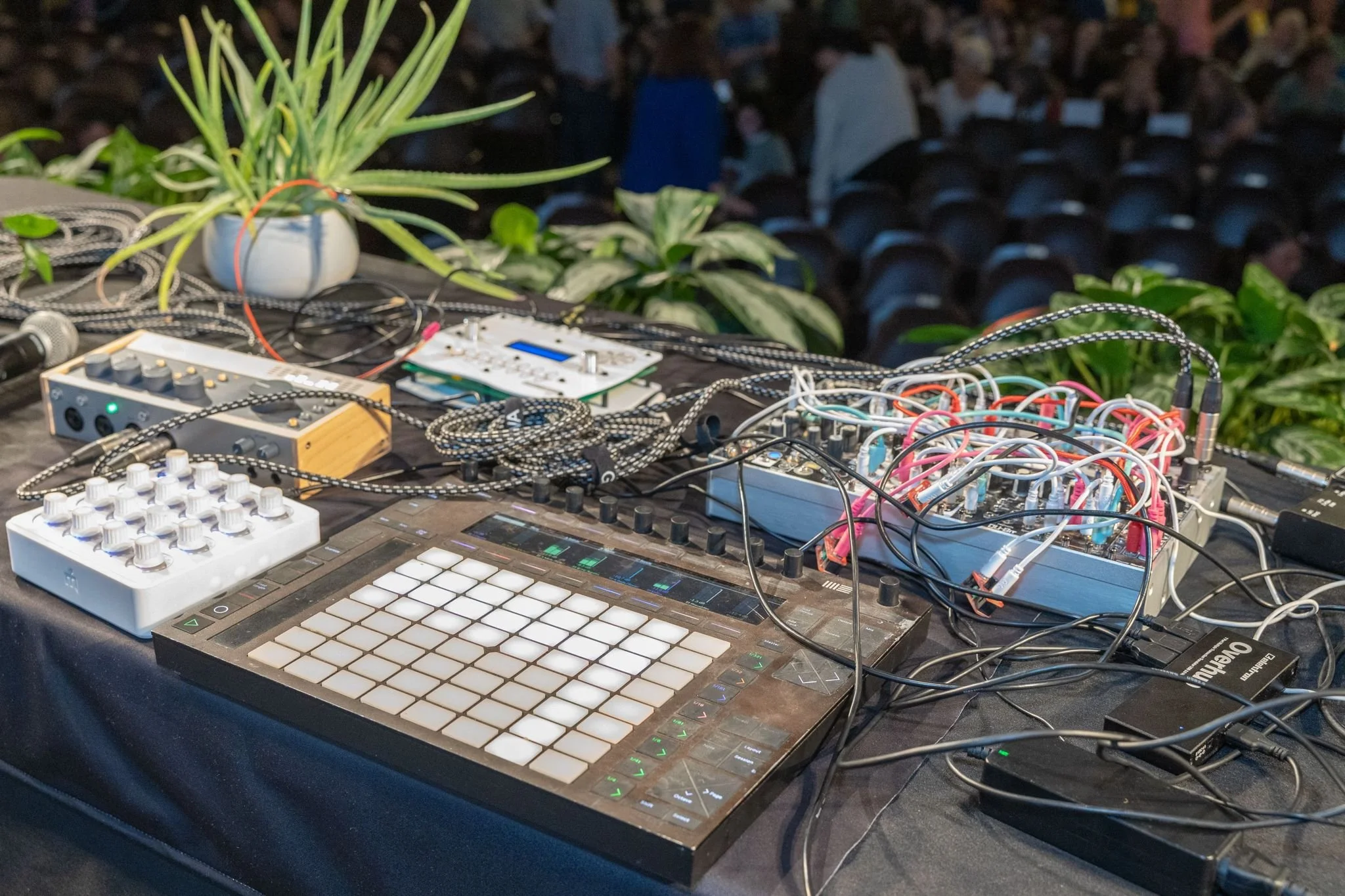 Electronic music equipment setup on a table, including a grid-based MIDI controller, synthesizers, mixers, connected with numerous cables, with potted plants in the background and an audience in an auditorium.