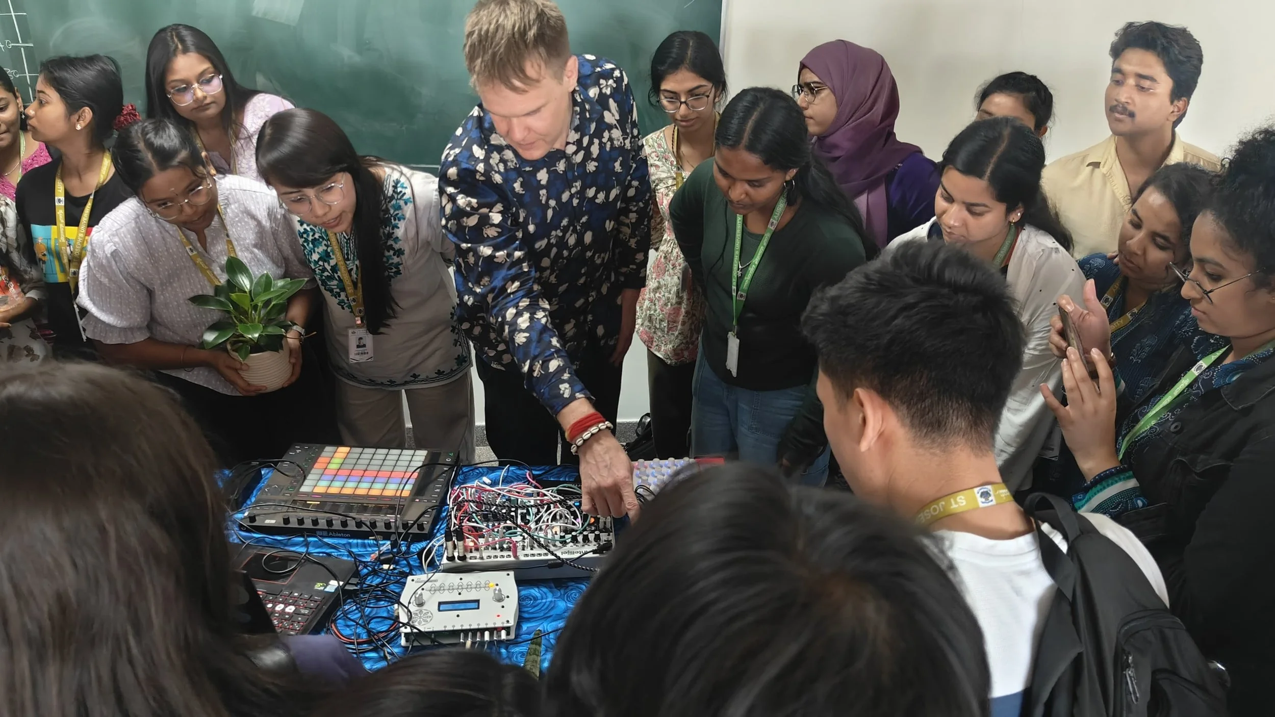 A diverse group of students and a teacher gathered around electronic music equipment in a classroom with a chalkboard in the background, observing and listening.