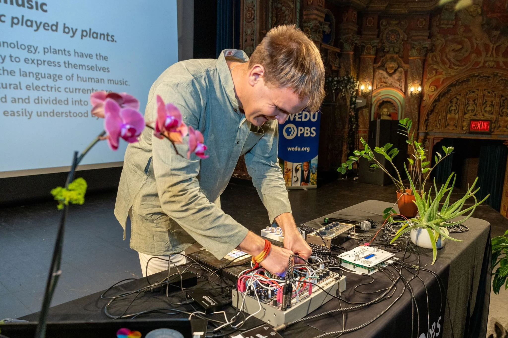 A man is performing with electronic music equipment on a table, in a decorated theater or venue, with orchids and plants on the table, and banners in the background.