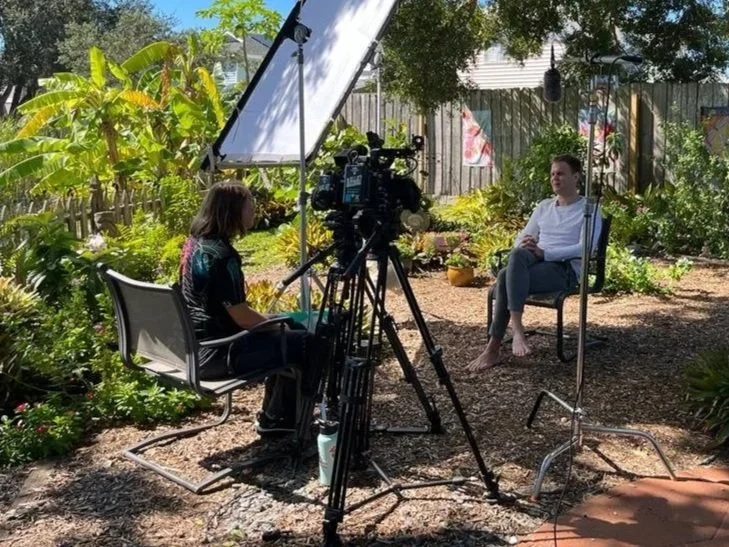 A woman conducting an outdoor interview with a man in a garden, with recording equipment and camera setup