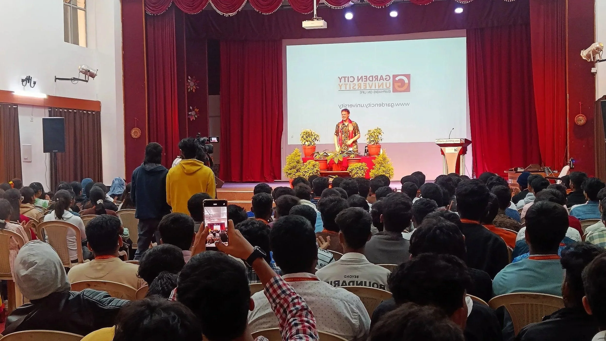 A speaker on stage at a conference or seminar, standing behind a table with potted plants, in front of a large screen displaying the logo and name of 'GardenCity University' in an auditorium filled with an audience.