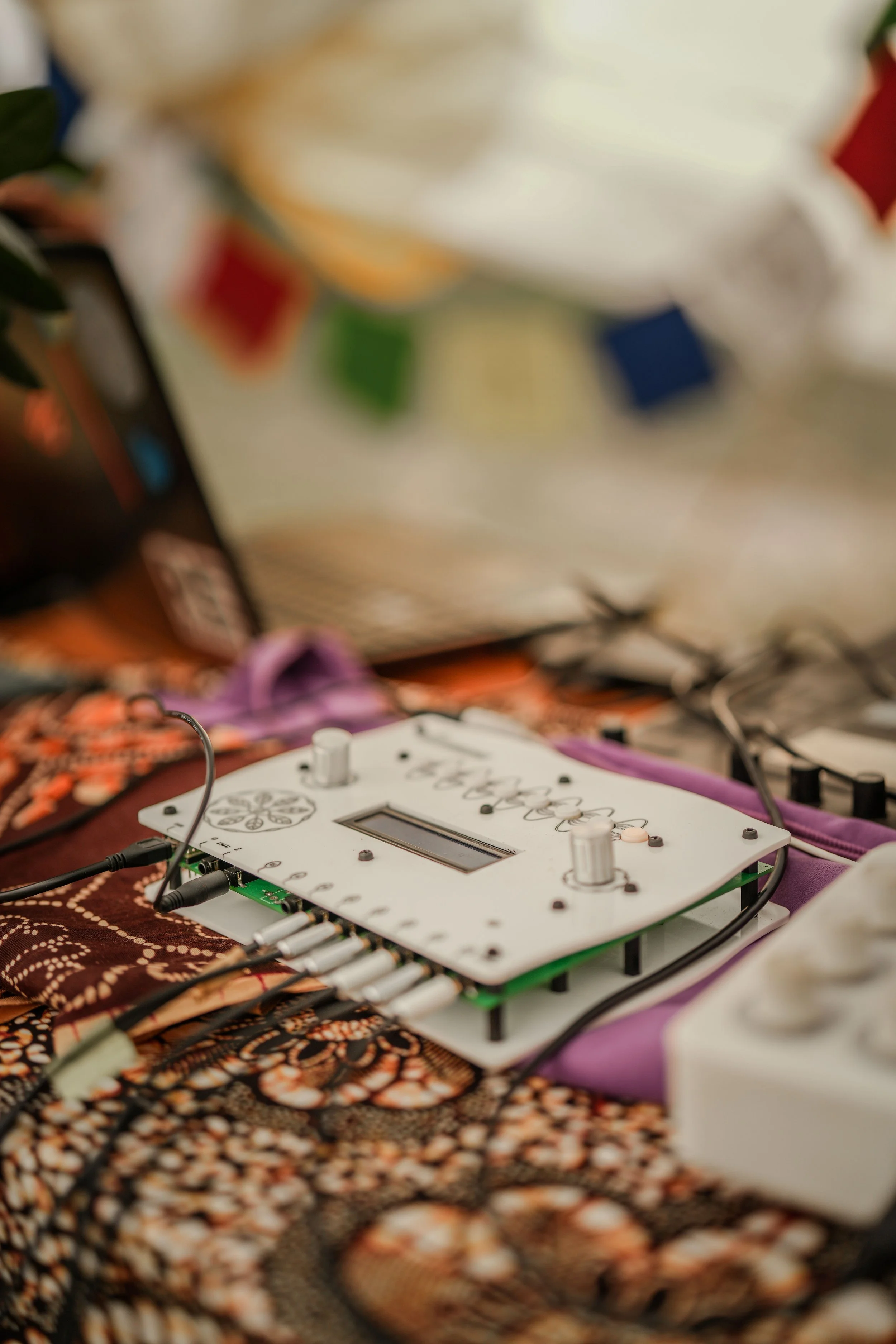 Electronic device with knobs, sliders, and a digital display placed on a patterned fabric surface, surrounded by cables.