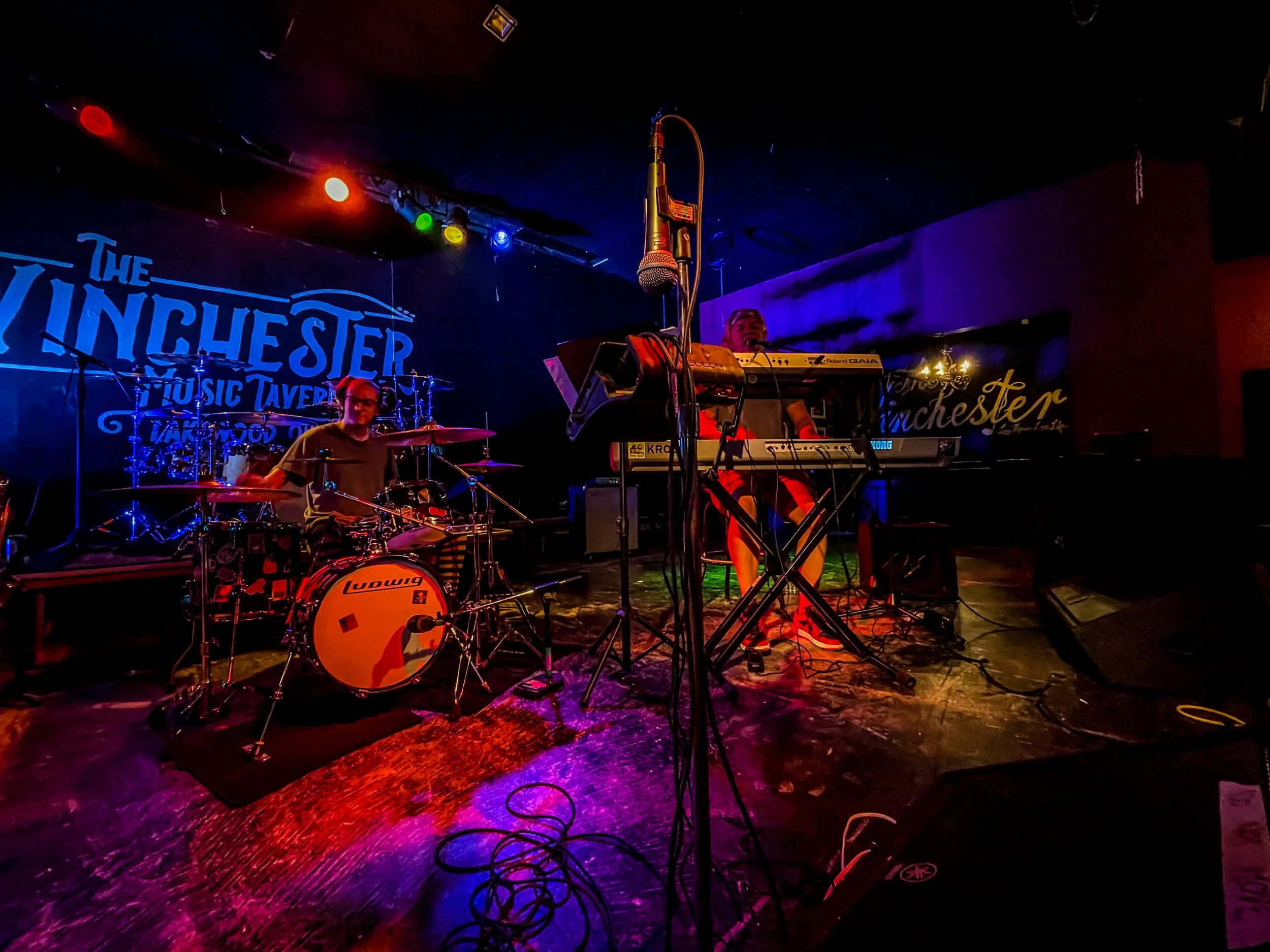 Musicians performing on stage at The Winchester Music Tavern with colorful stage lighting, drum set, keyboard, and microphones.