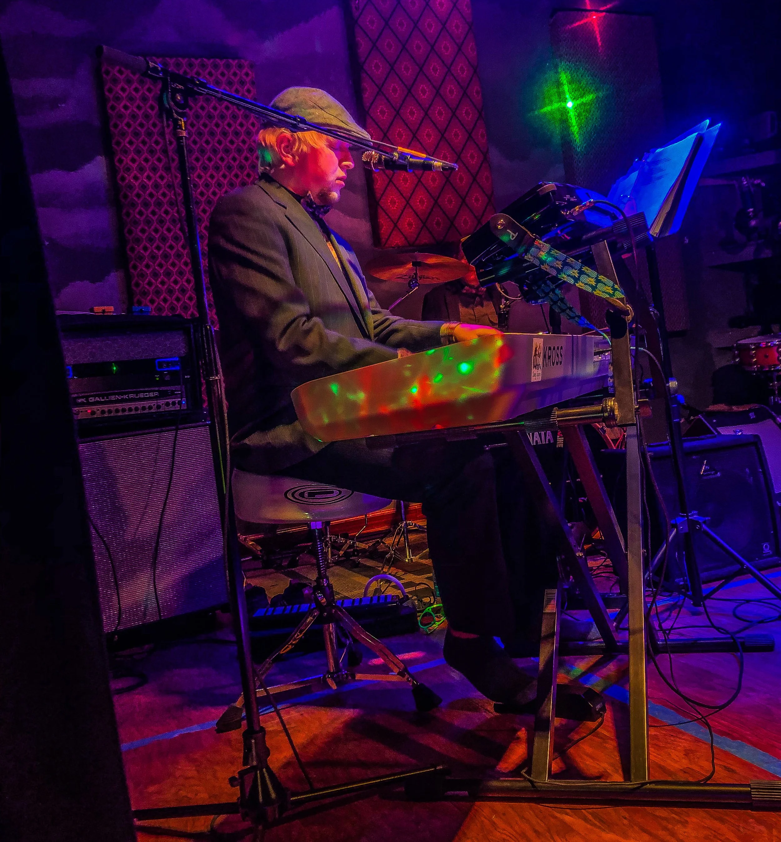 A man in a suit playing a keyboard on stage with colorful lights and sound equipment around him.