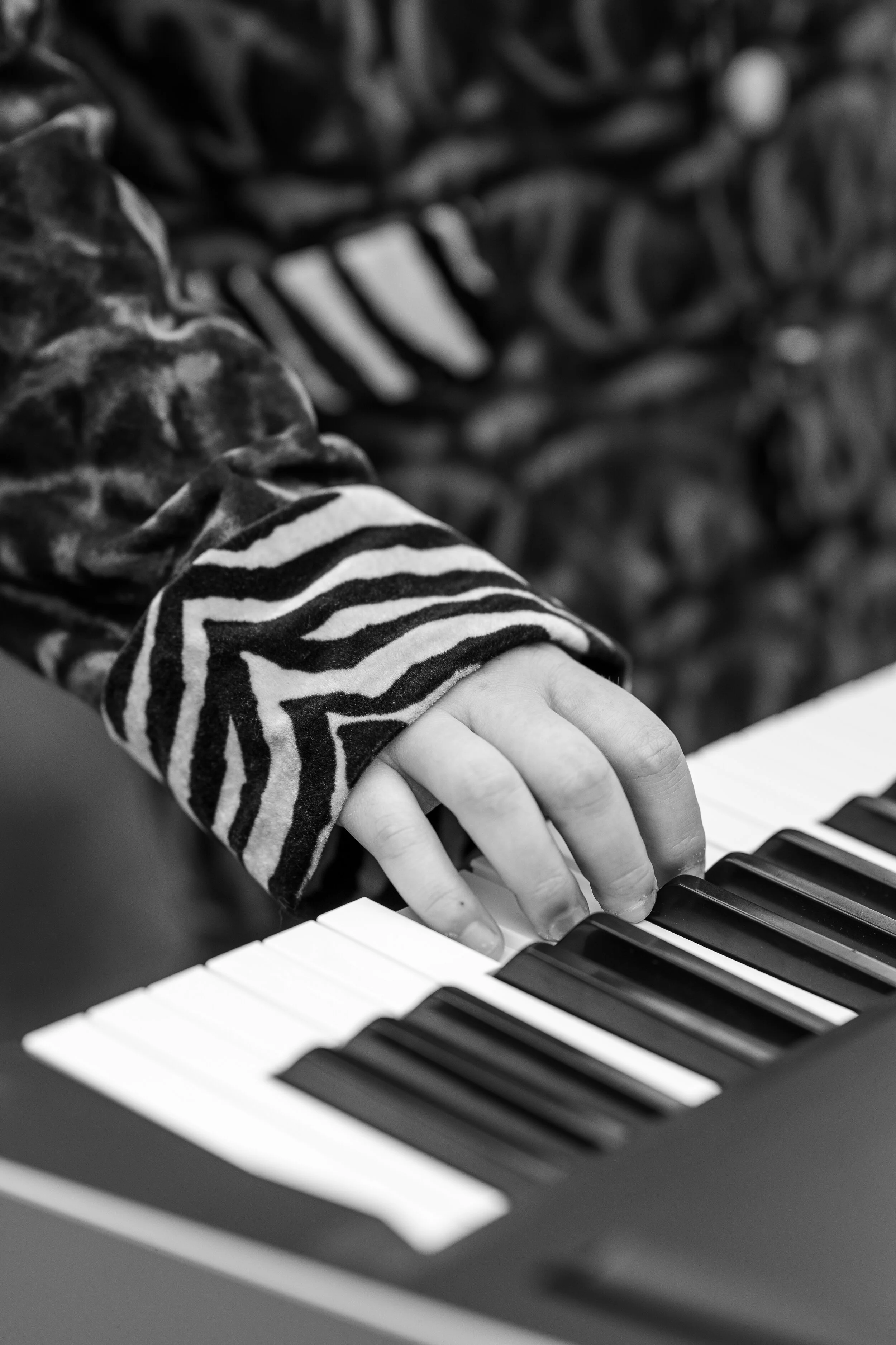 Close-up of a child's hand playing a piano, wearing a camouflage jacket with zebra-striped cuffs, in black and white.