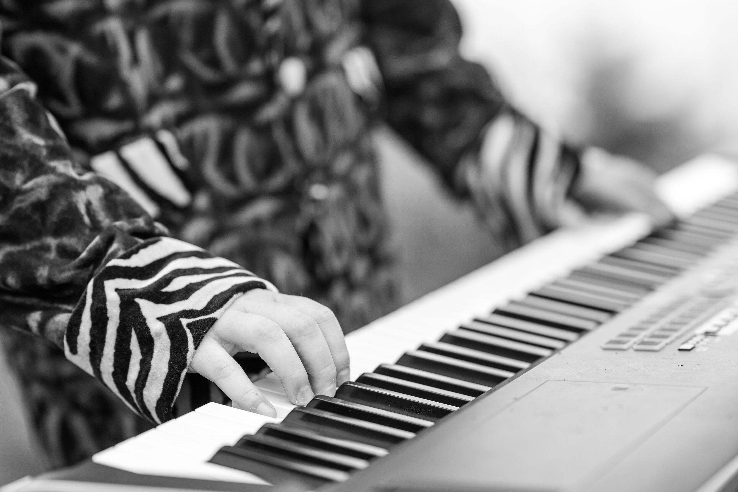 A child's hand pressing piano keys, wearing a long-sleeved shirt with zebra stripe pattern, in black and white.
