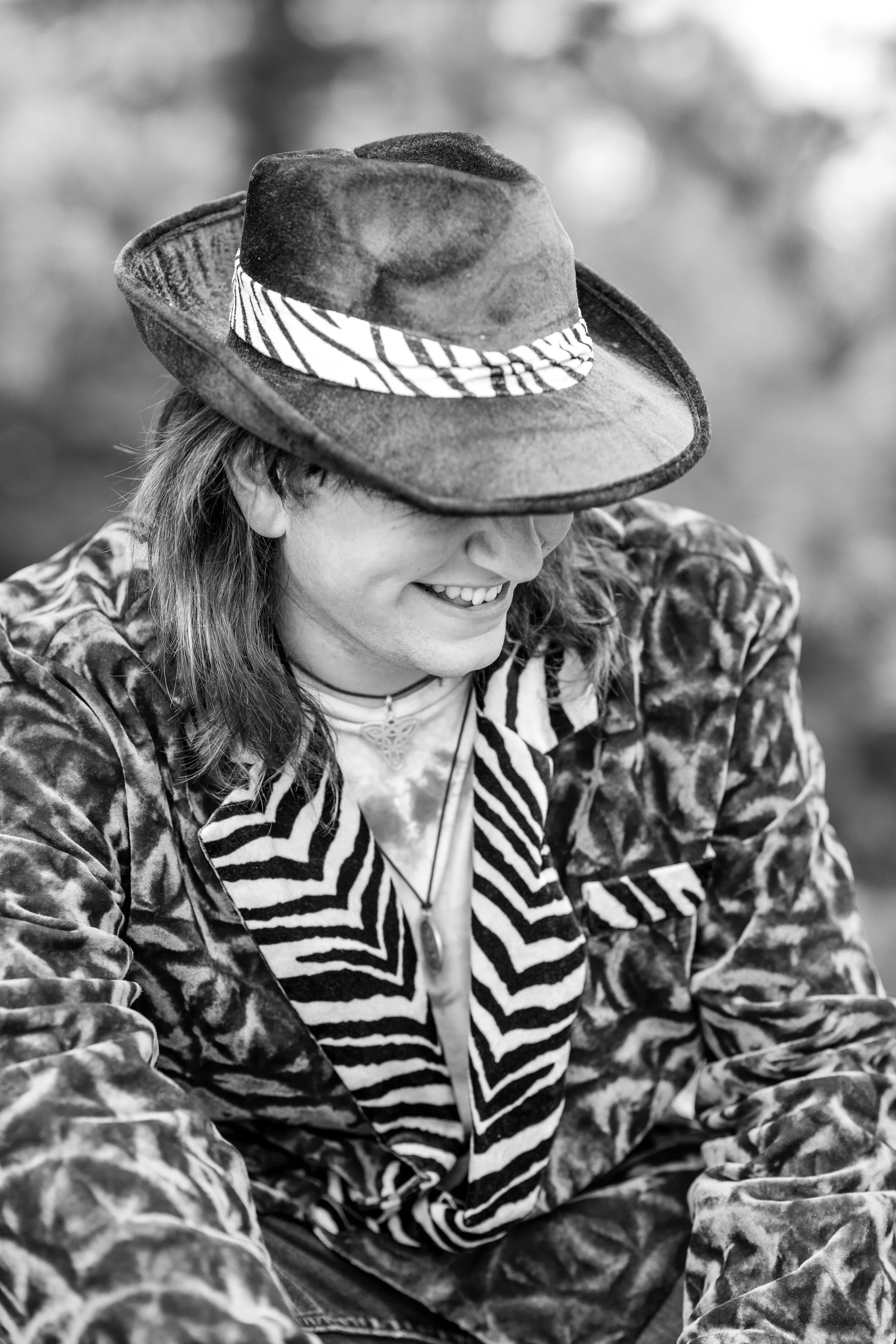 A person smiling, wearing a cowboy hat, patterned jacket, and zebra-striped scarf, in black and white.