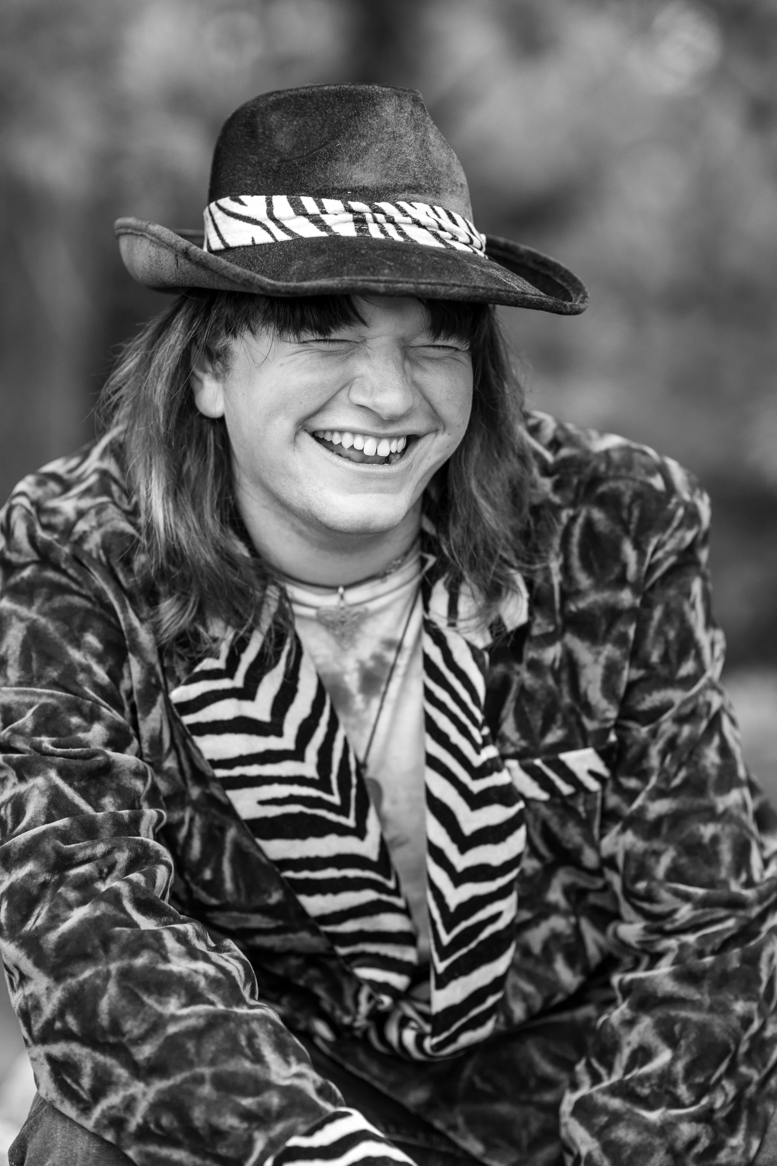 A person with long hair smiling widely, wearing a hat and a patterned jacket, captured in black and white.