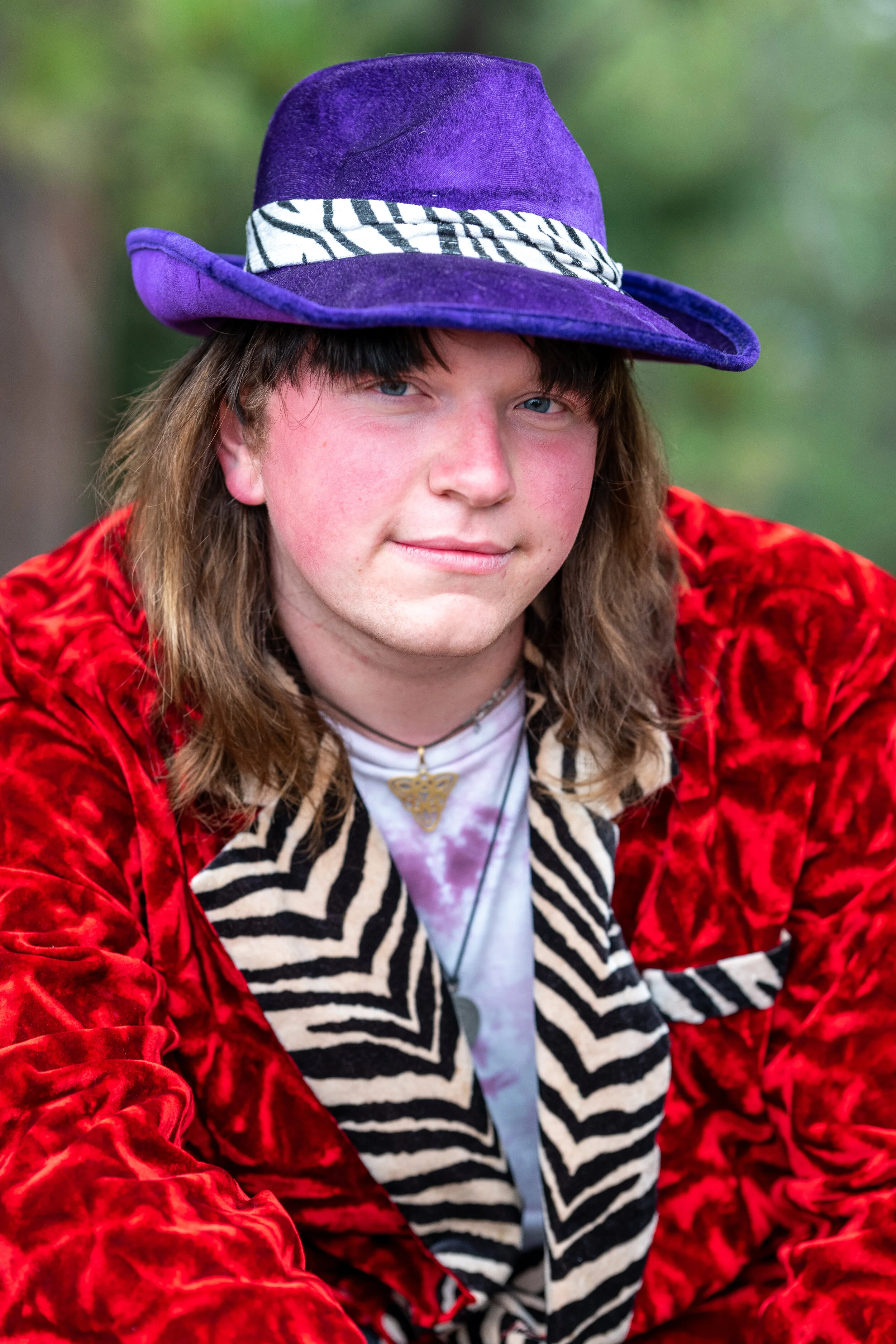A young person wearing a purple hat, red velvet jacket, zebra-patterned scarf, and layered necklaces, outdoors with blurred green background.