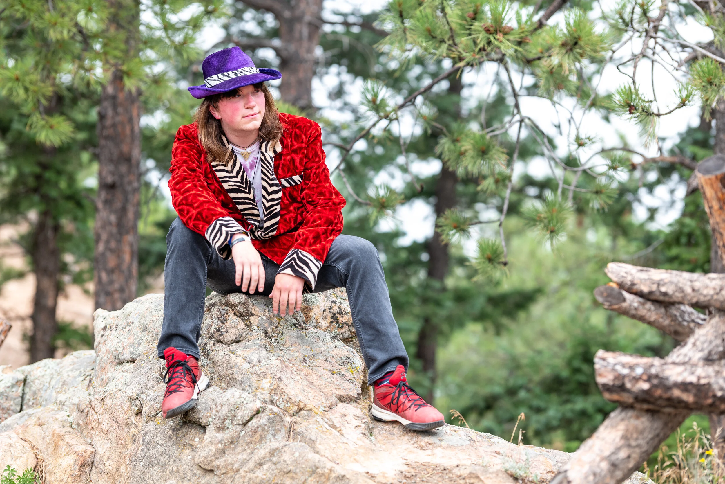 A person sitting on a large rock outdoors in a forest, wearing a purple hat, red velvet jacket with zebra print accents, black jeans, and red hiking shoes.