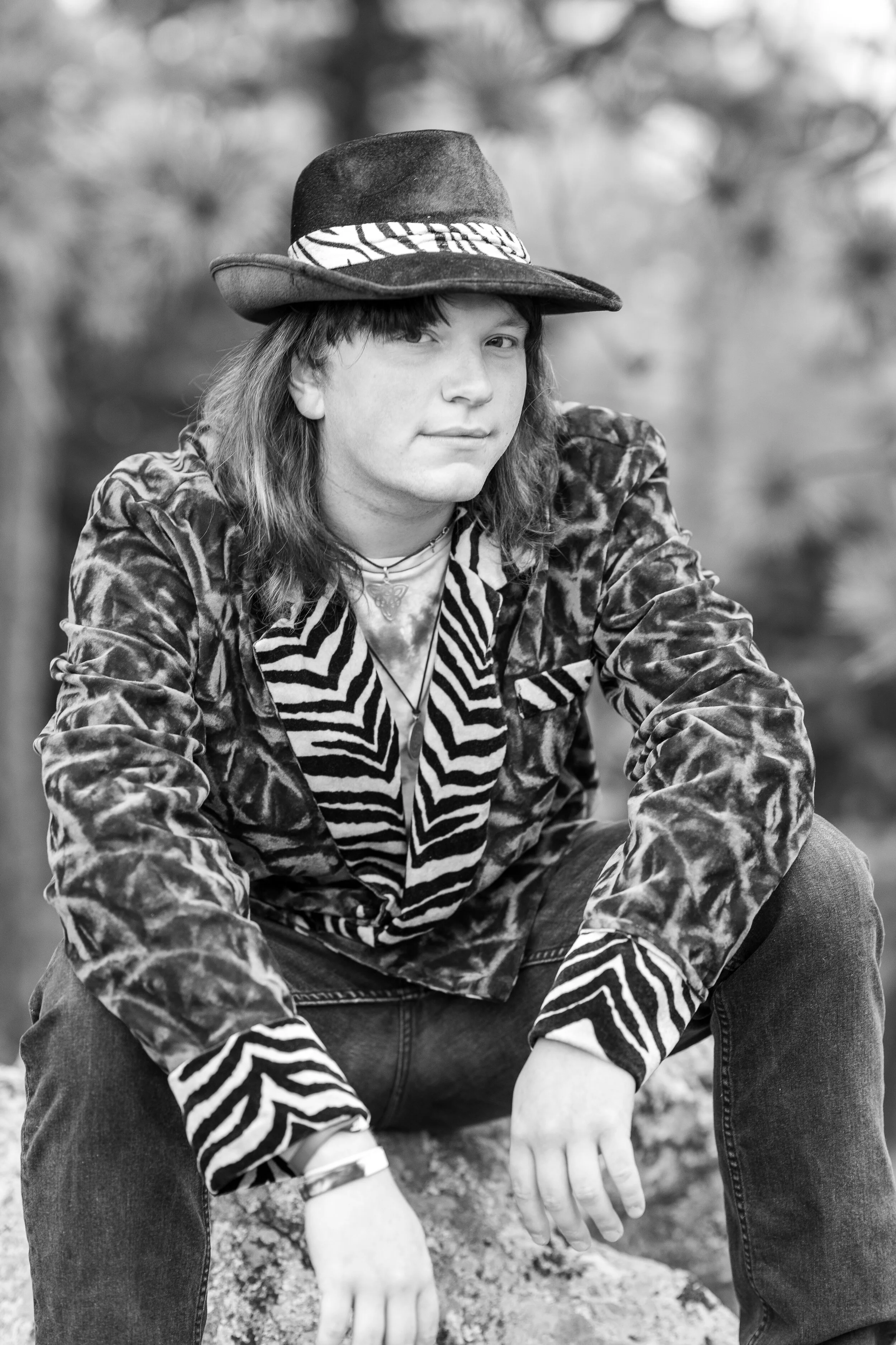 Black and white photo of a young man wearing a patterned hat and a velvet jacket with animal print details, sitting outdoors with a blurred natural background.