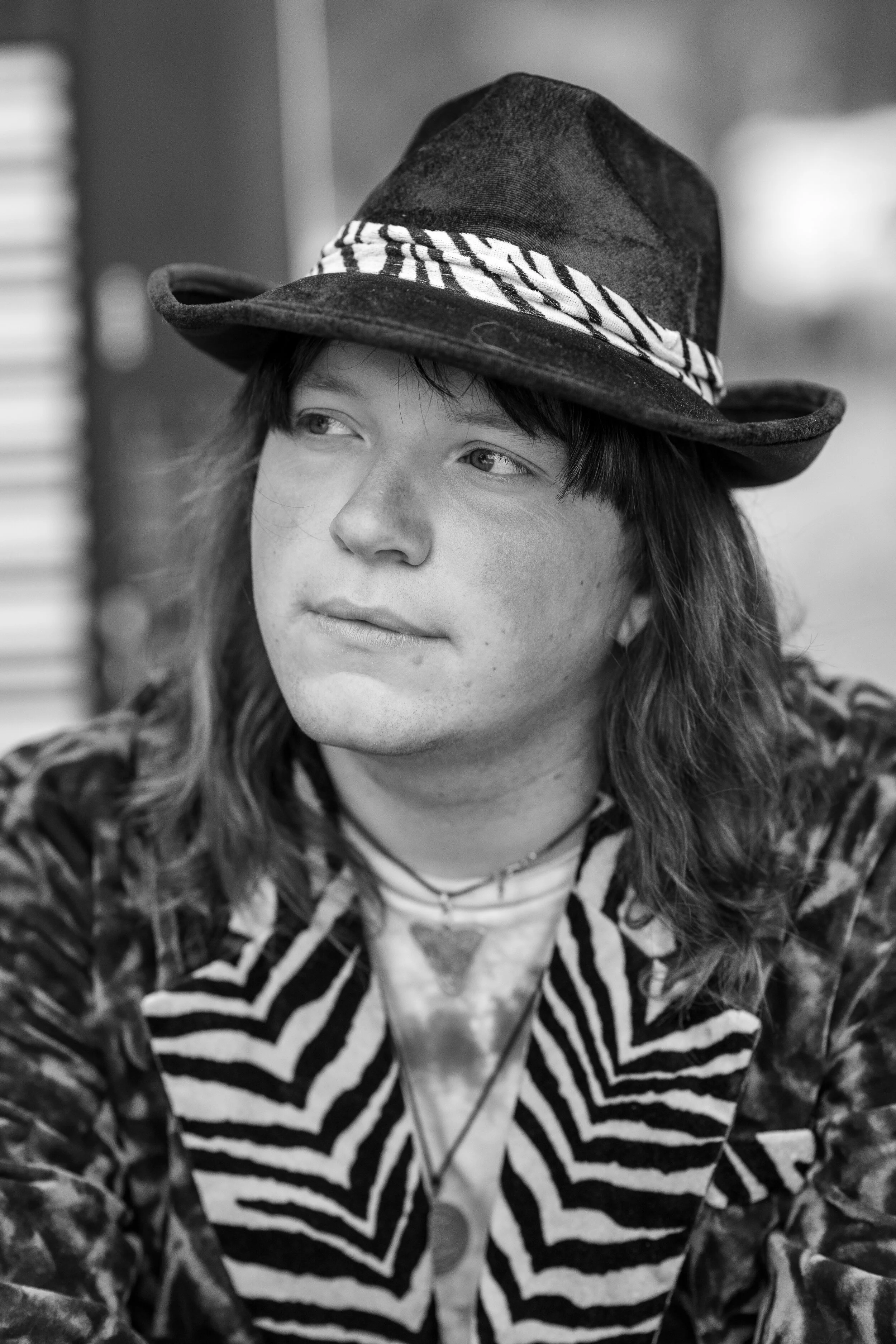 A young man with long hair wearing a wide-brimmed hat, patterned jacket, and necklaces, looking to the side in a black and white photo.