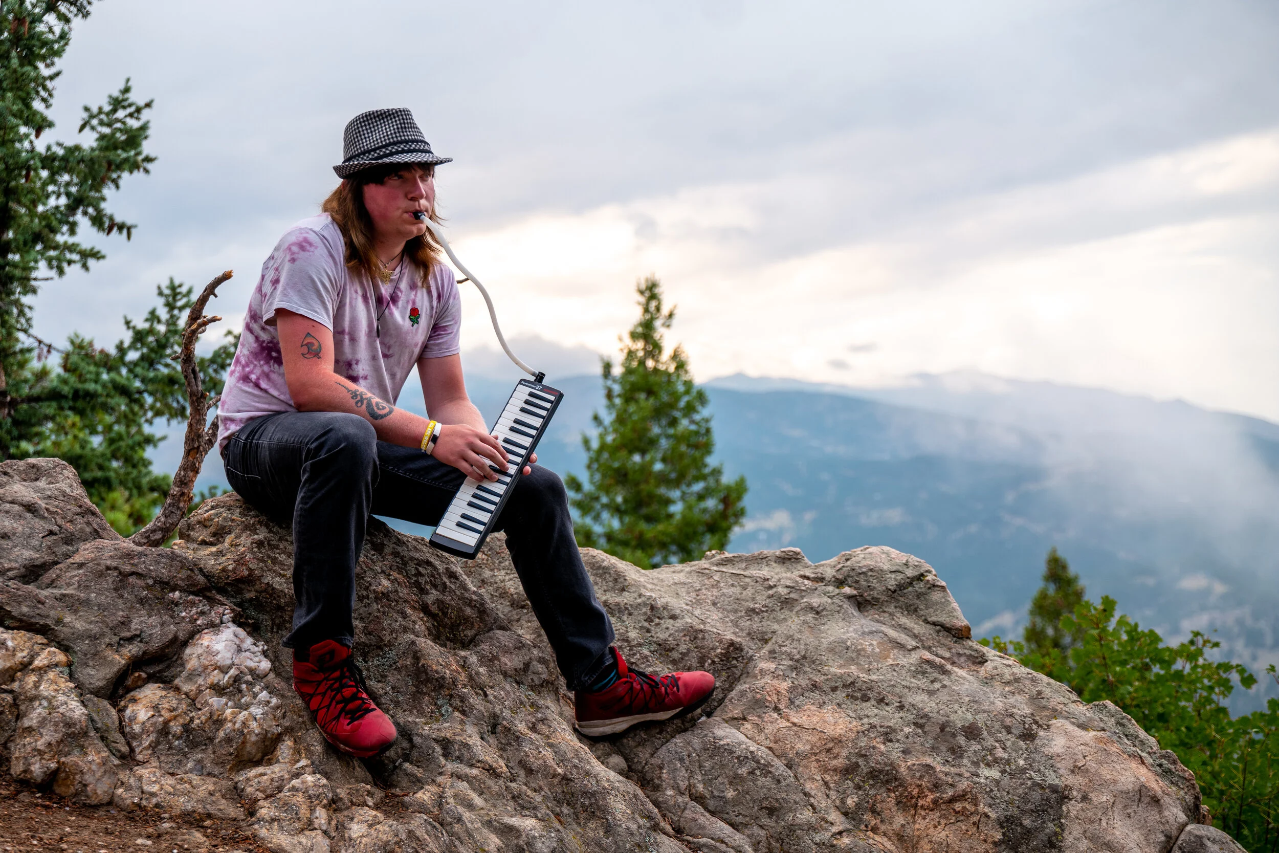 A person sitting on rocks outdoors, playing a keytar and singing into a microphone, with a mountain landscape and trees in the background.