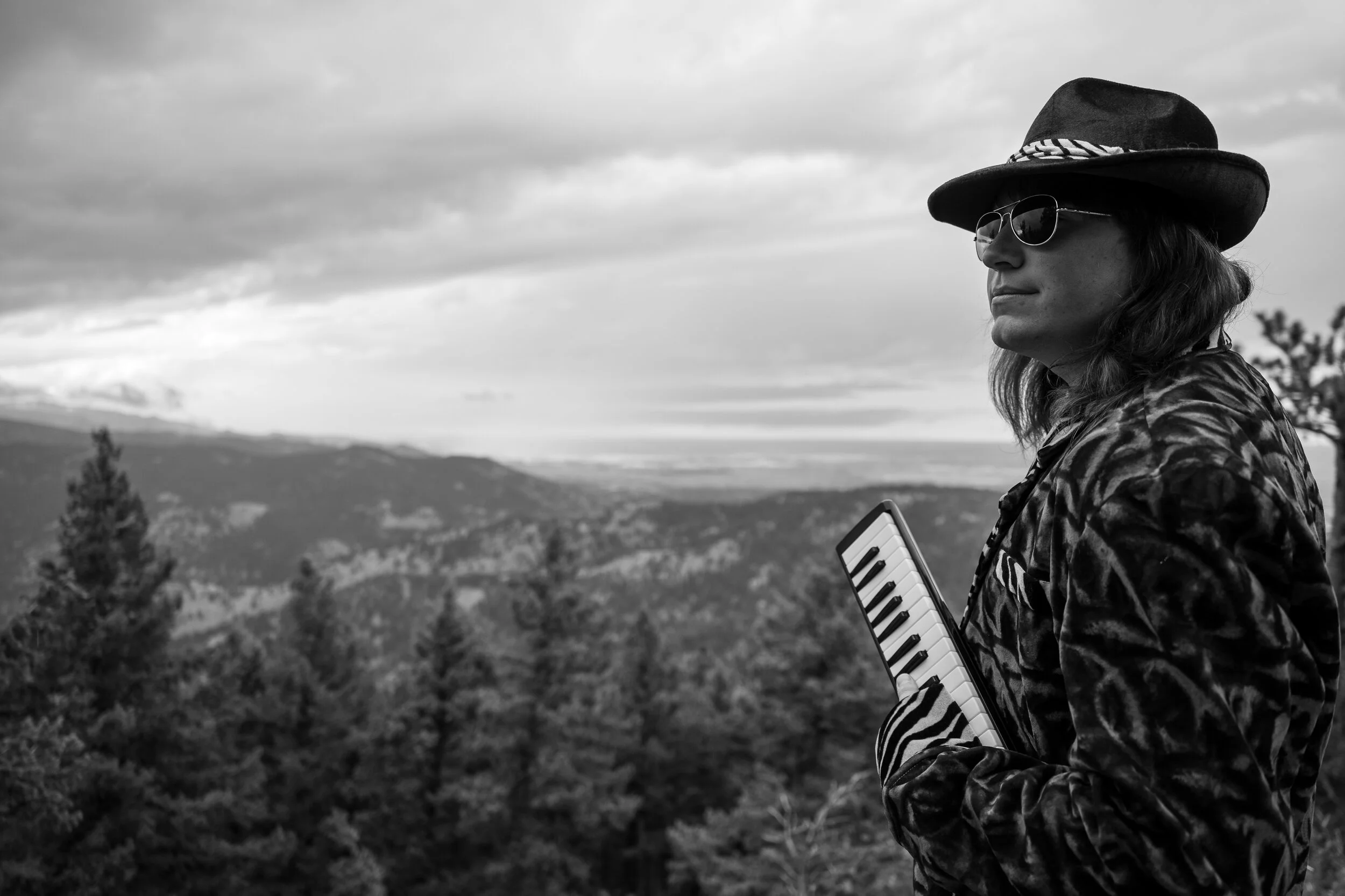 A person wearing sunglasses and a hat, holding a keyboard, standing outdoors in a mountainous landscape with trees and clouds in the background.