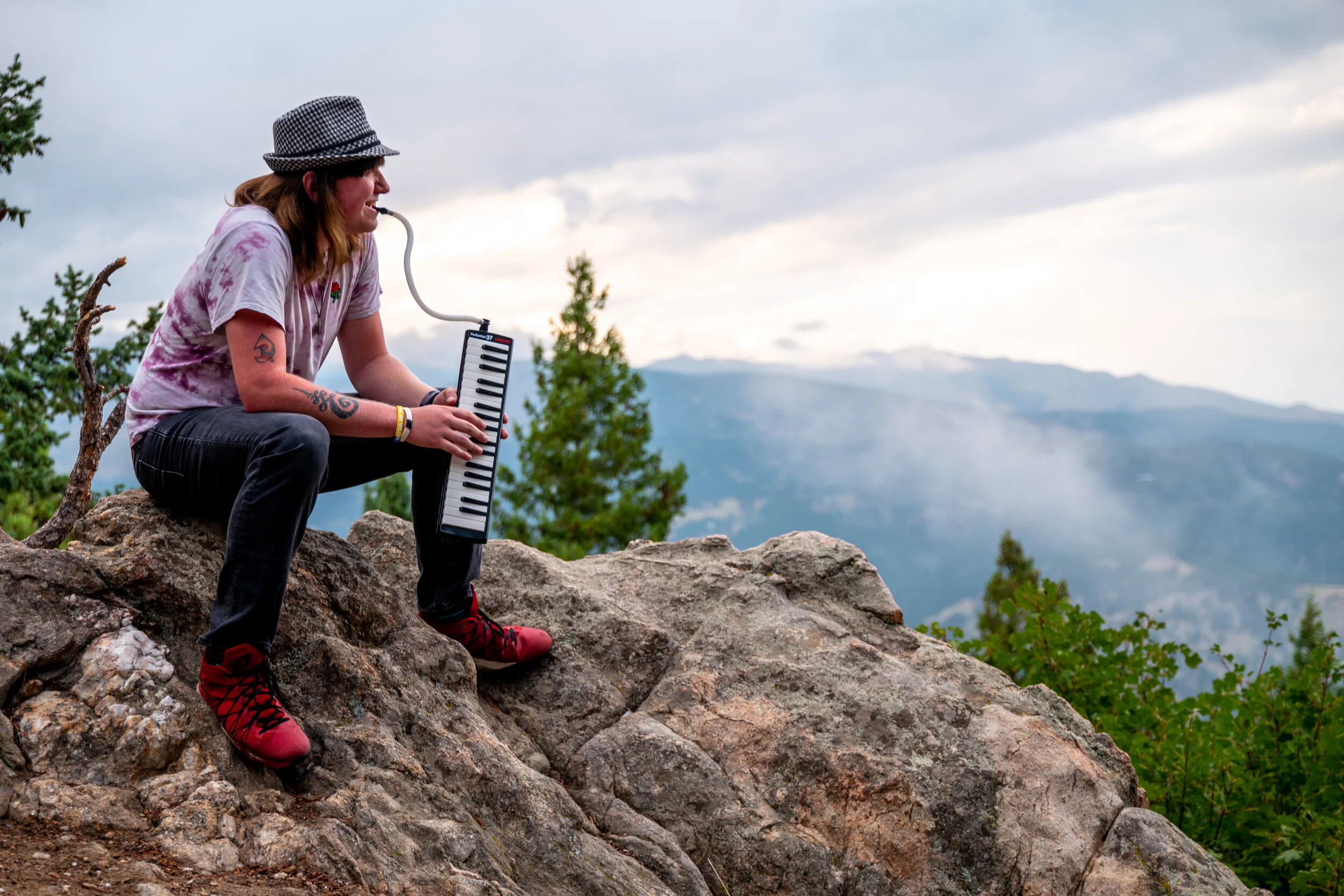 A woman sitting on rocks outdoors, playing a small electronic keyboard with a mouthpiece, surrounded by mountains, trees, and a cloudy sky.