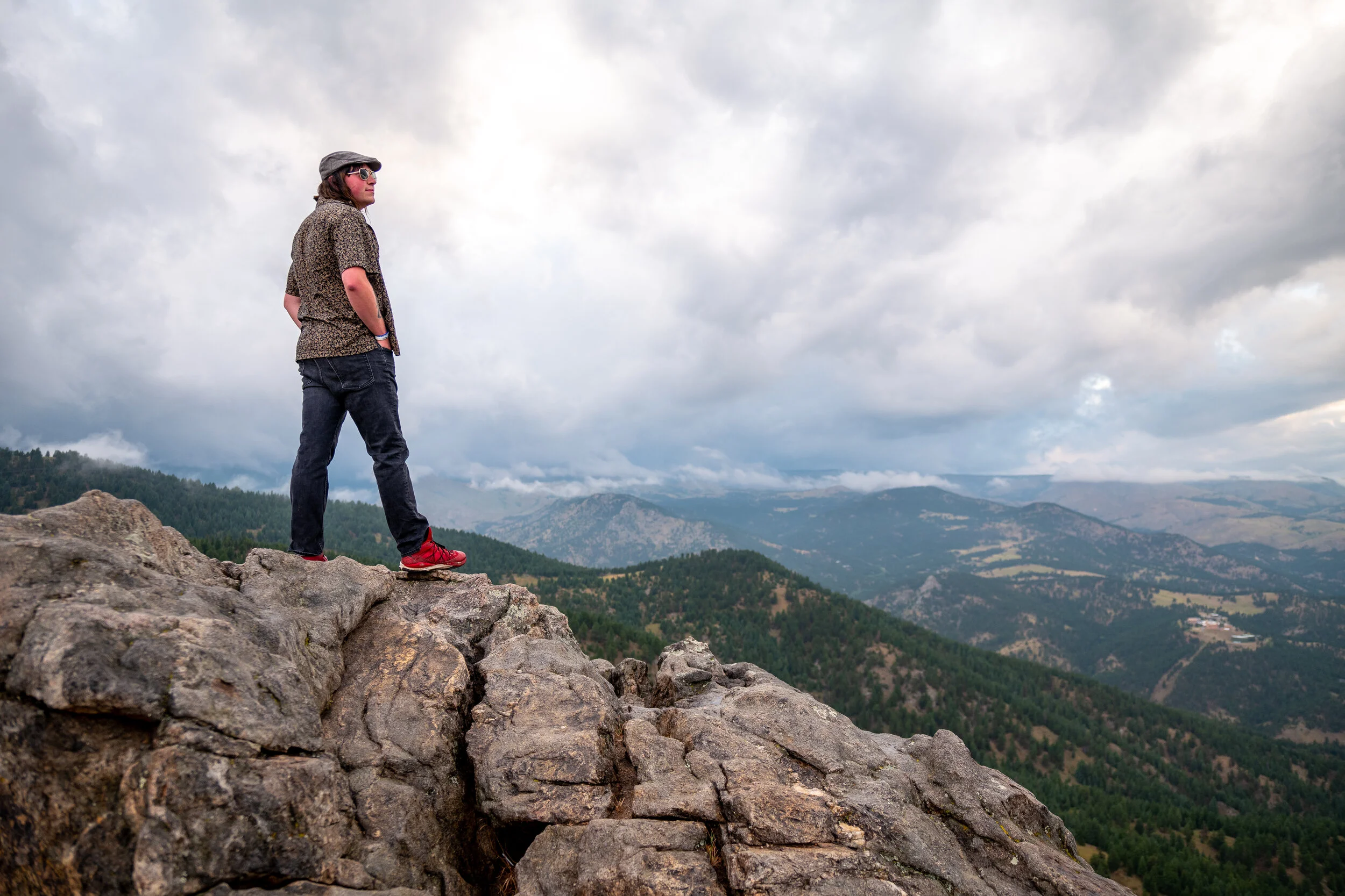 Person standing on rocky ledge overlooking a mountainous landscape with clouds in the sky.