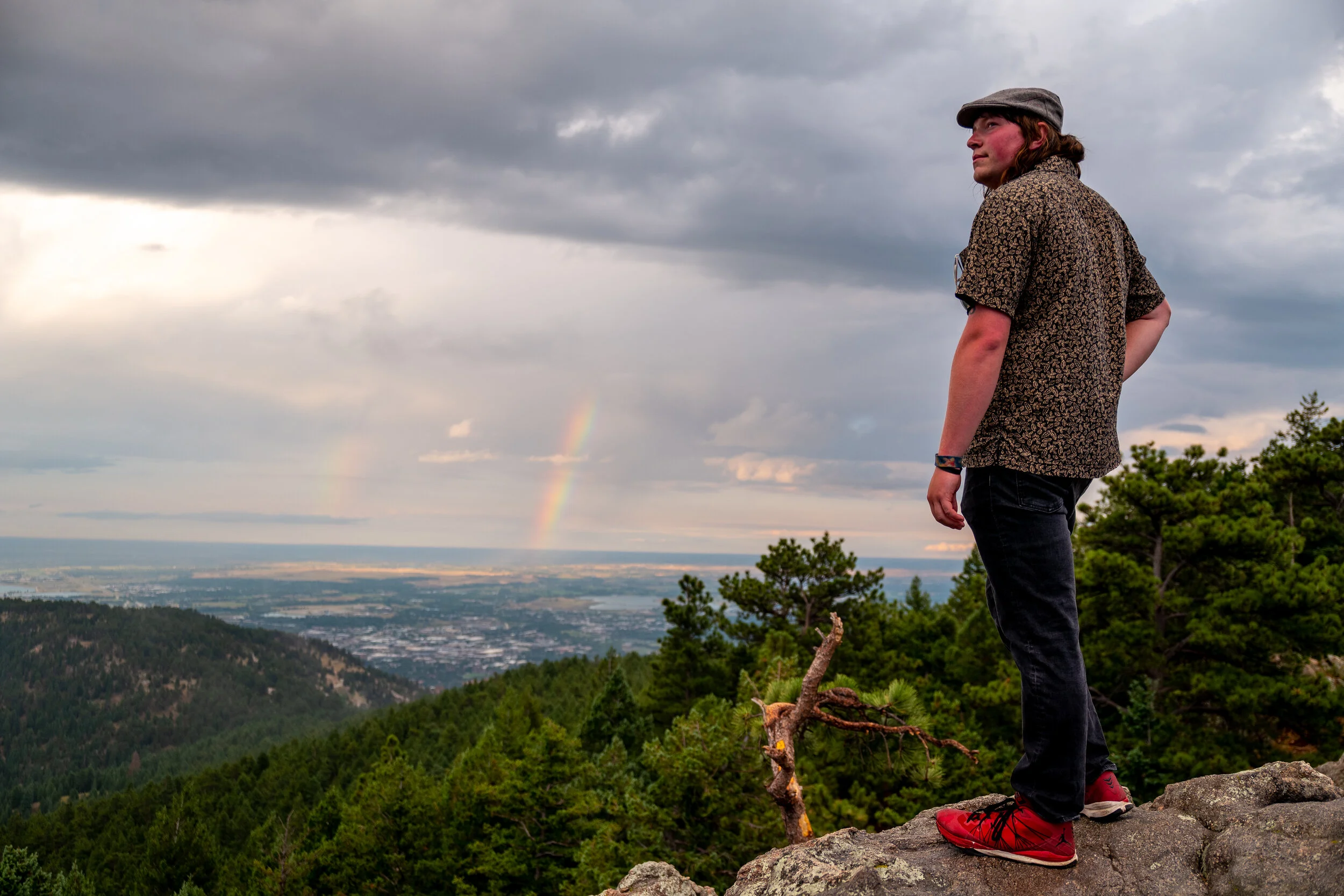 A man standing on a rocky outcrop on a mountain, looking to the side, with a view of a forest, city, and a rainbow in the distance under a cloudy sky.