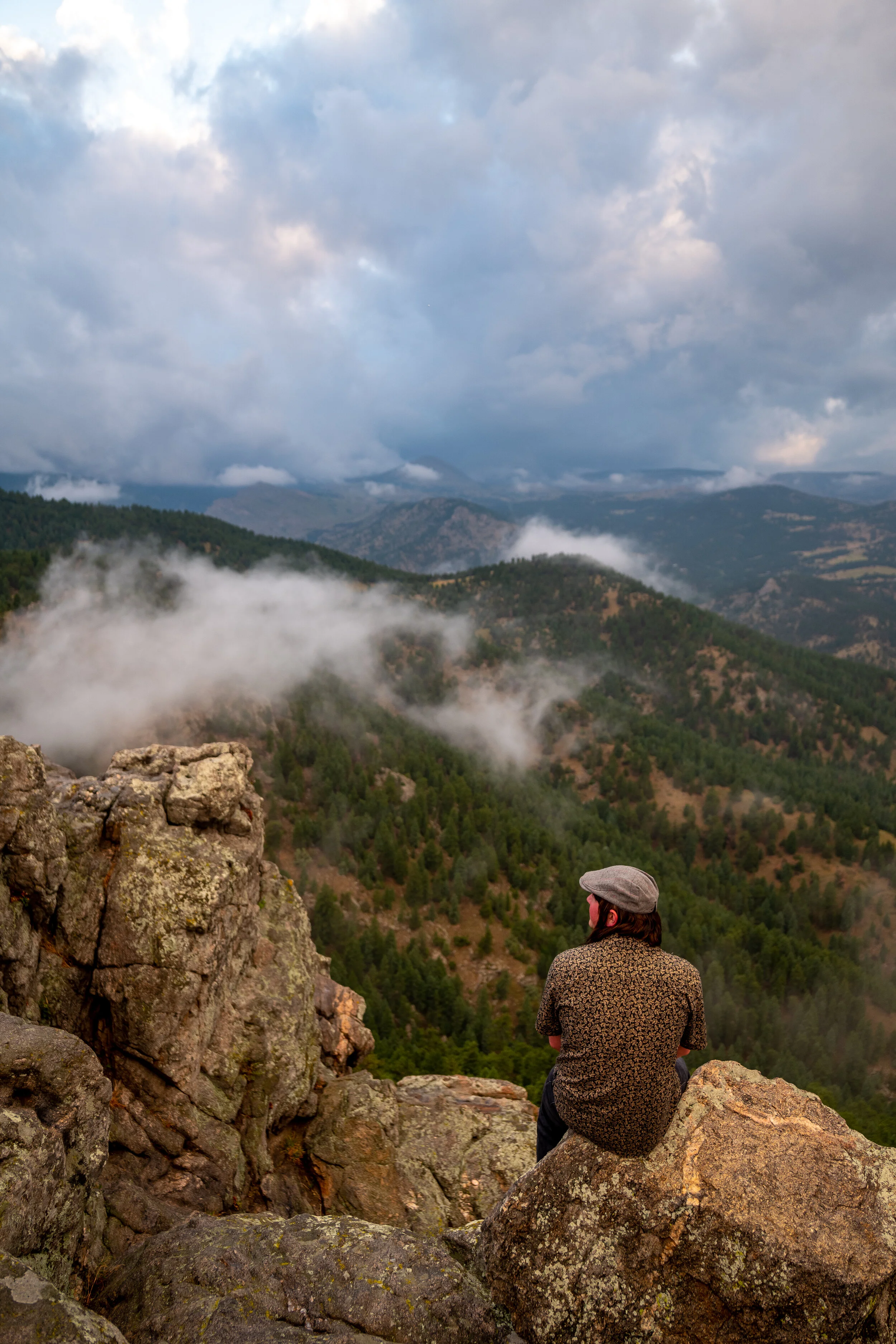 A person wearing a flat cap and patterned shirt sitting on rocks, looking out over a mountainous landscape with trees, clouds, and mist.