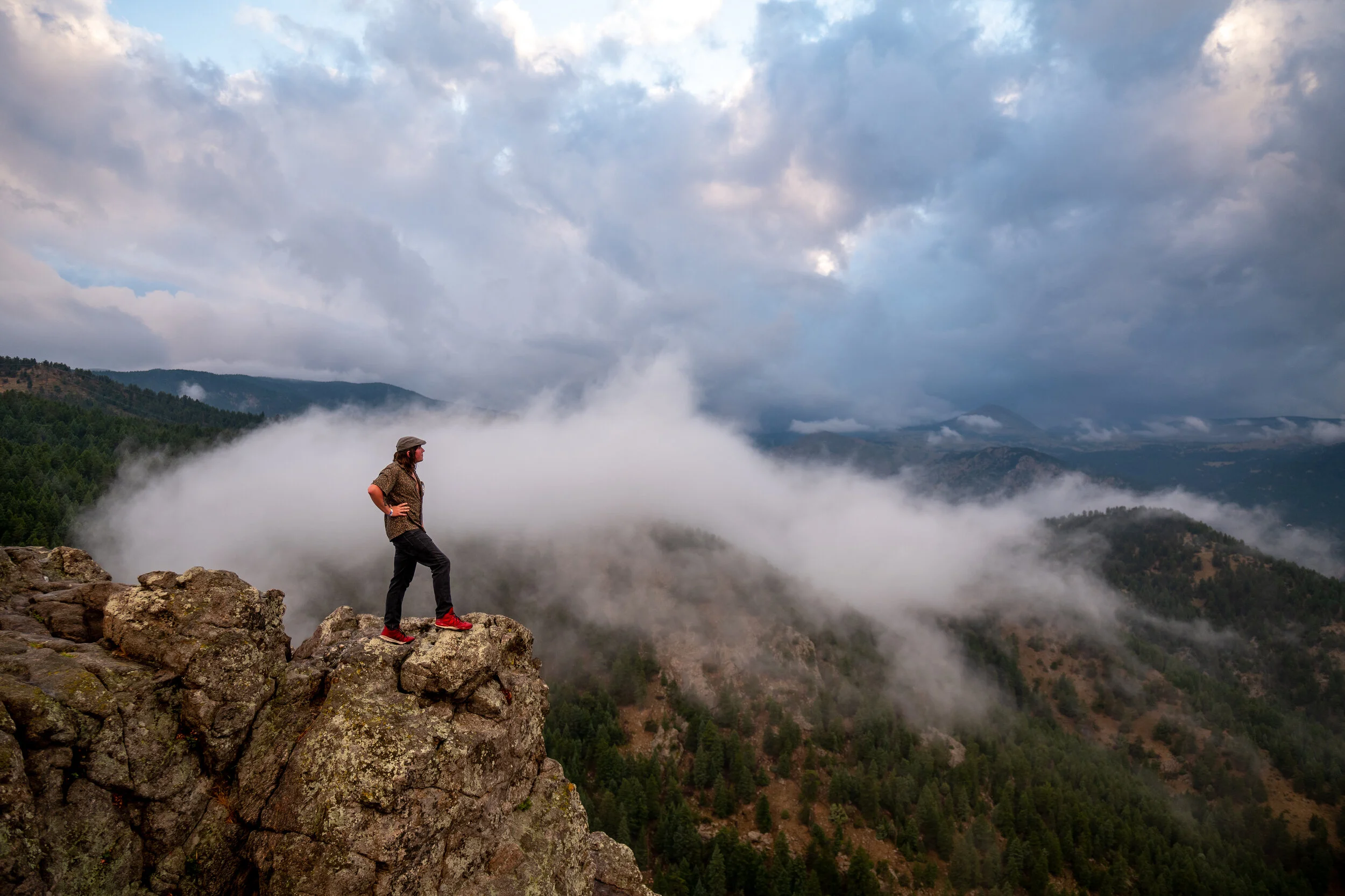 A person standing on a rocky cliff overlooking a mountainous landscape with fog, trees, and cloudy sky.