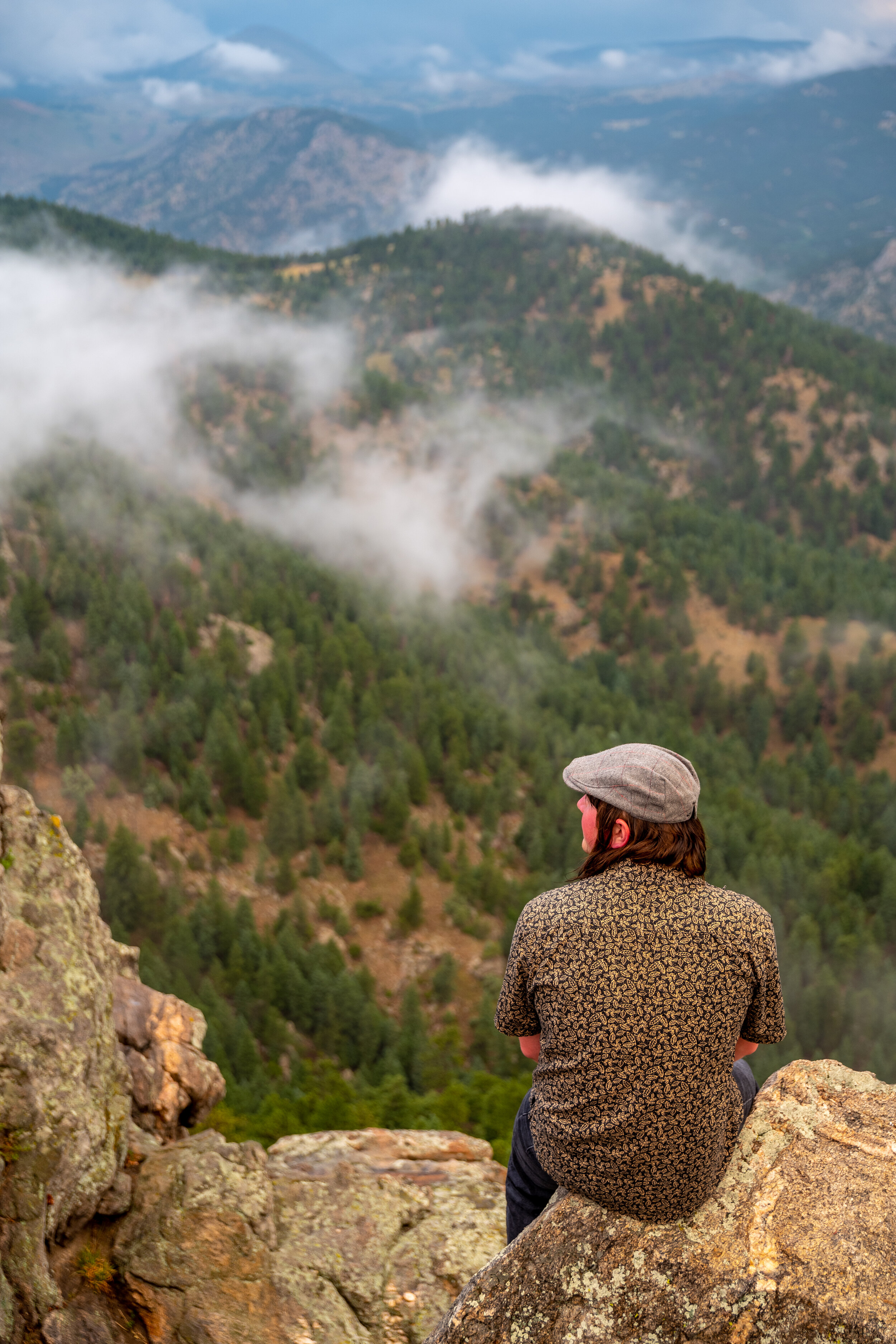 Person wearing a patterned shirt and gray hat sitting on a large rock, overlooking a forested mountain landscape with mist or clouds.