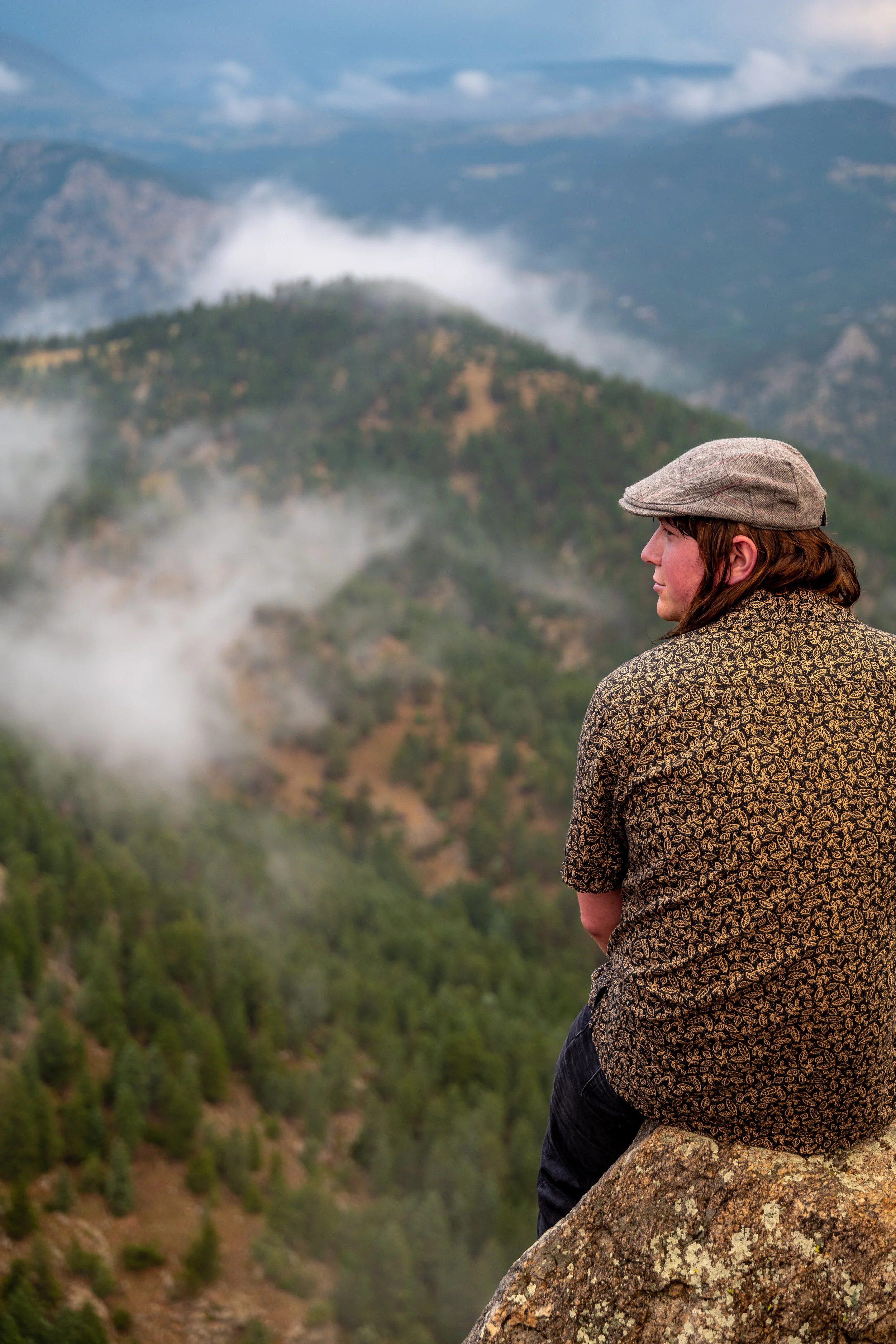 A woman sitting on a rocky ledge overlooking a mountainous landscape covered in trees and clouds.