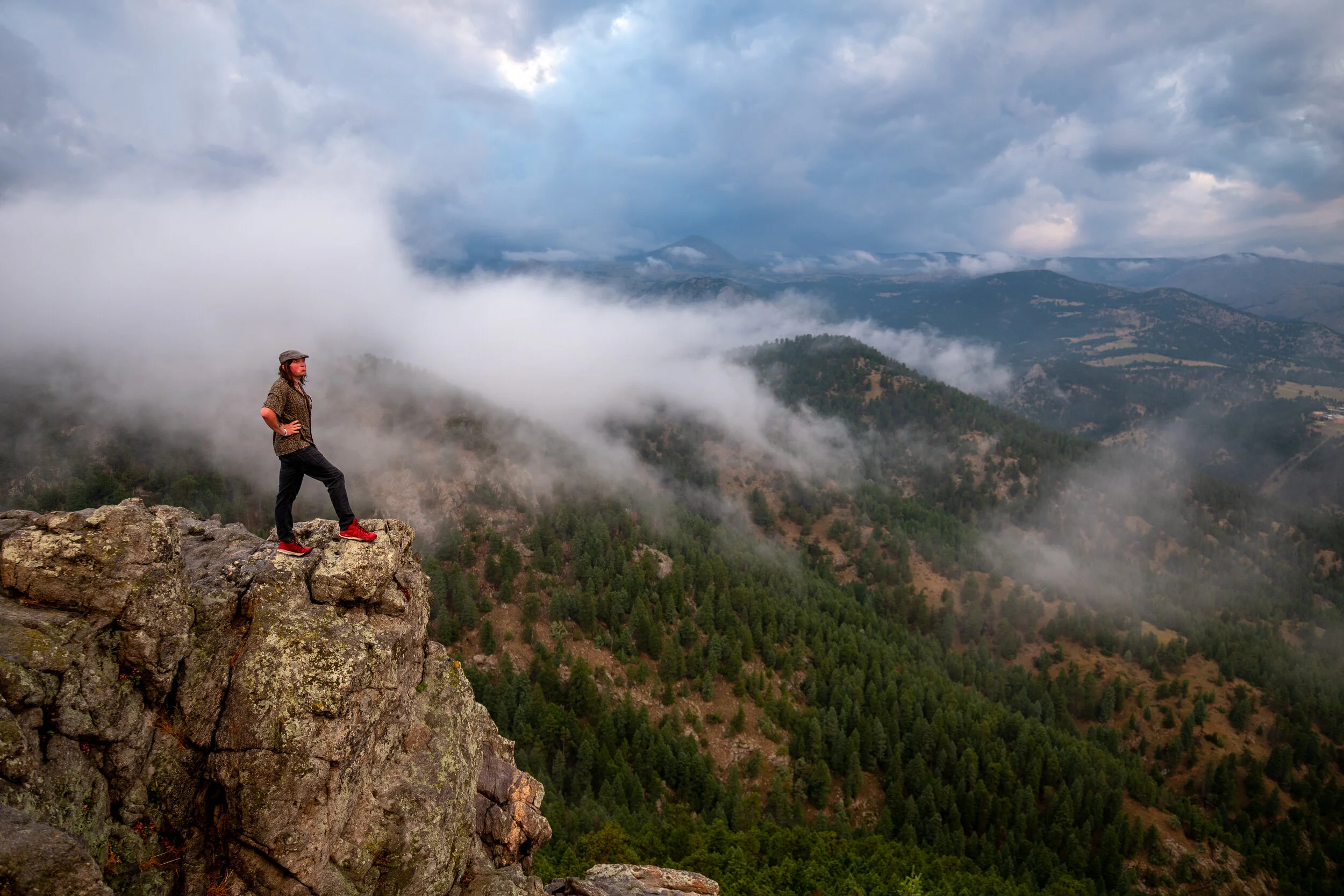 A person standing on a rocky cliff overlooking a foggy mountainous landscape with trees and clouds.