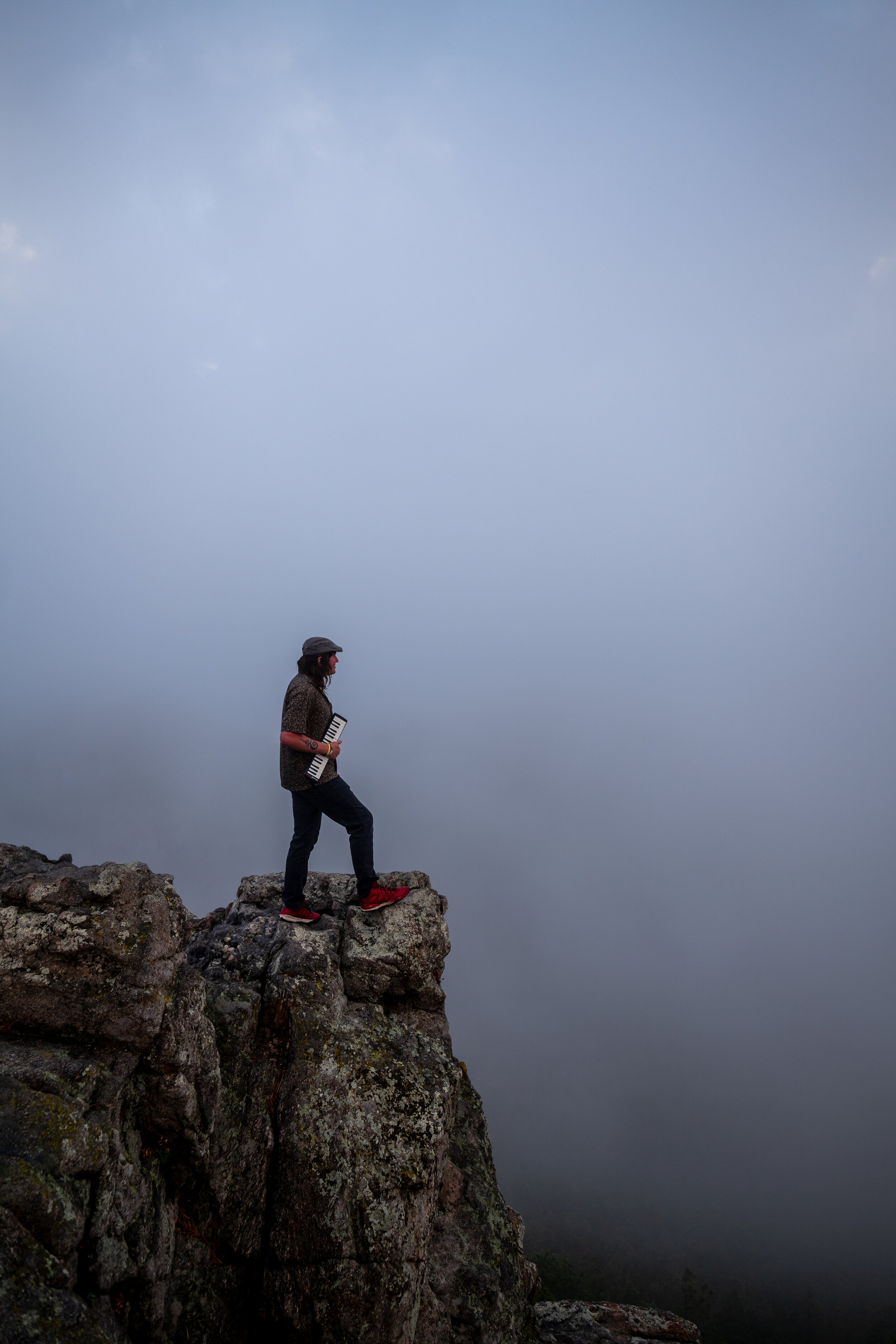 Person standing on a rocky ledge at the edge of a cliff, holding a small keyboard, with a cloudy sky and fog below.