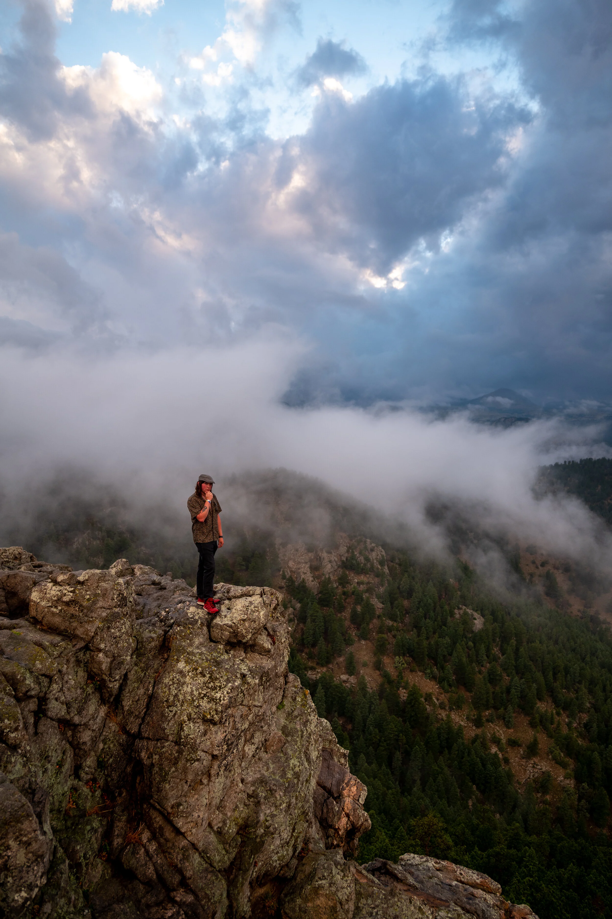 A person standing on a rocky edge on a mountain with fog, clouds, and forested hills in the background.