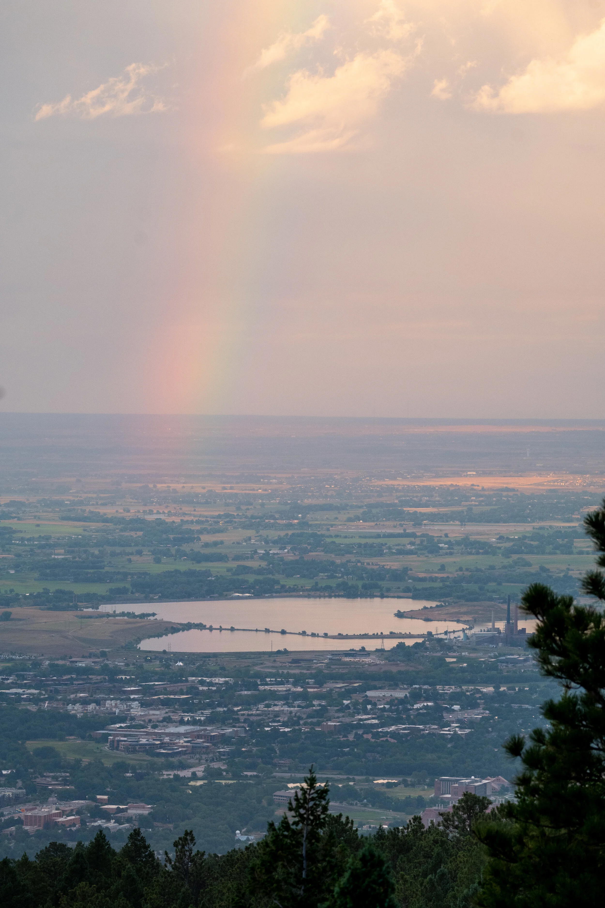 Scenic view of a city with a lake, trees in the foreground, and a rainbow in the sky with clouds.