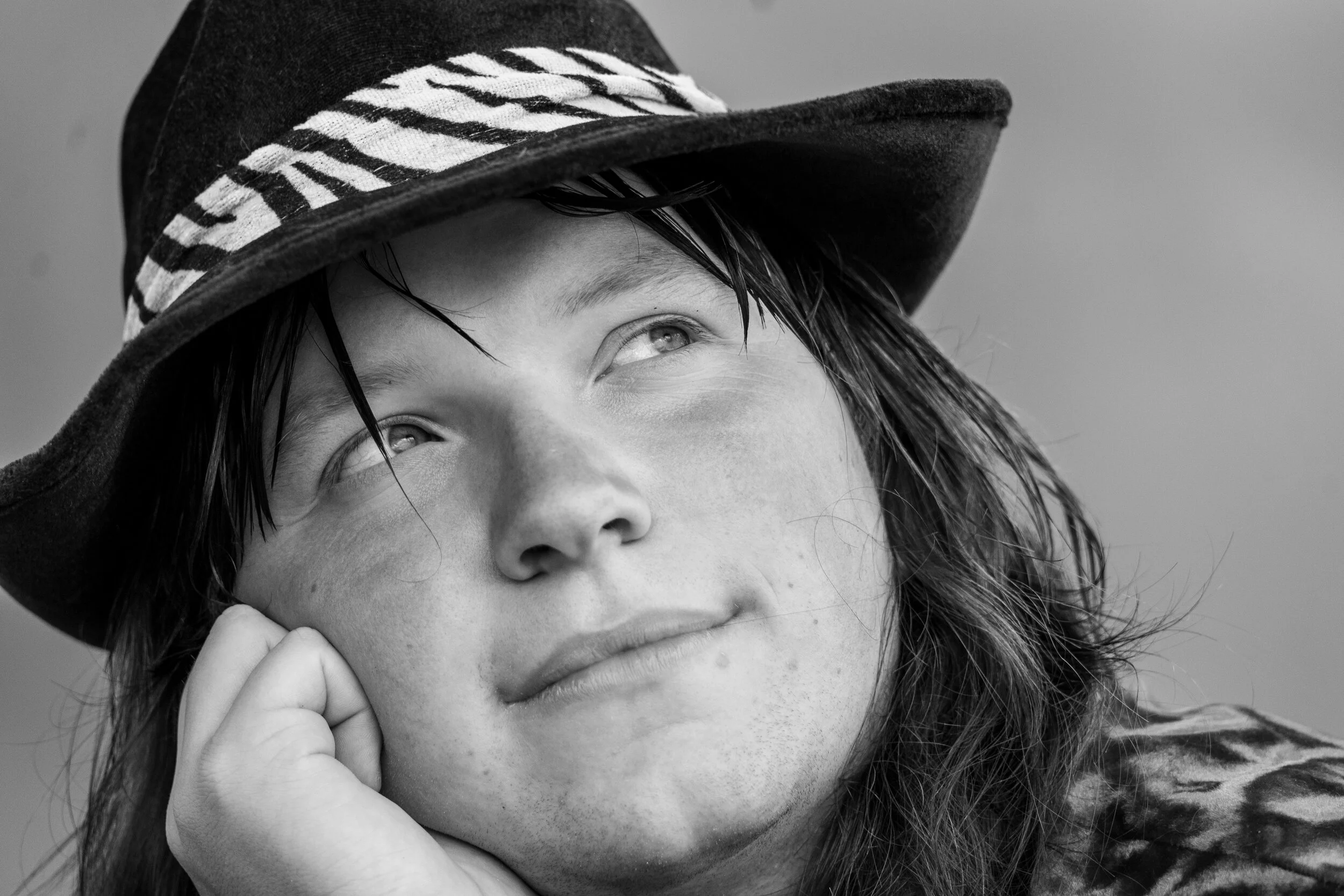 Close-up black and white portrait of a young woman wearing a wide-brimmed hat with a striped band, resting her face on her hand, looking upwards.