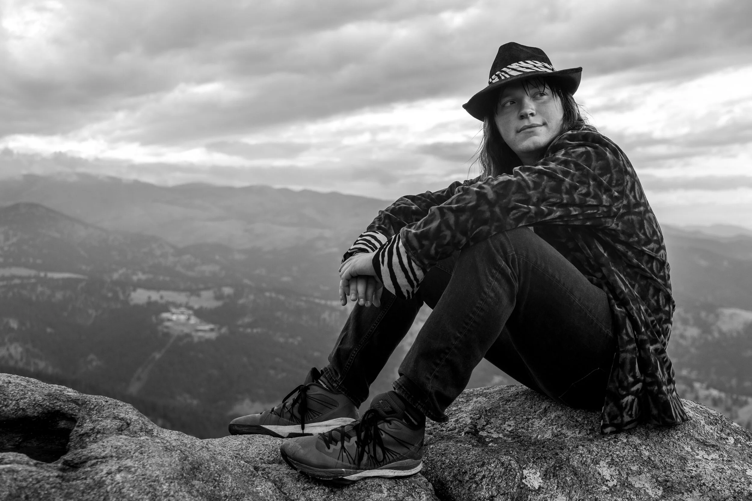 A person sitting on a large rock outdoors overlooking a mountainous landscape, wearing a hat, long sleeve shirt with animal print, and hiking shoes, with a cloudy sky above.