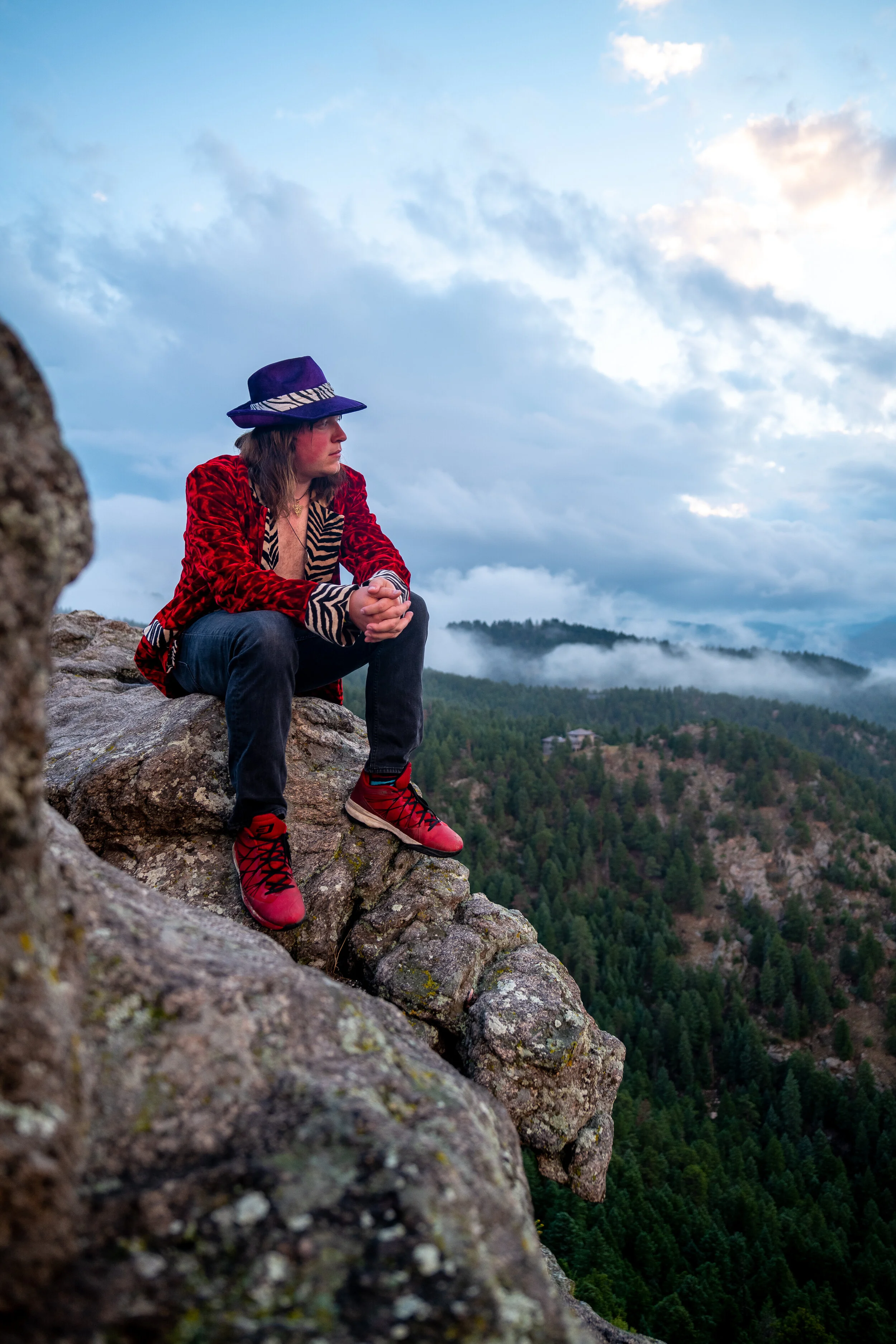 A woman sitting on a rocky ledge overlooking a forested mountain landscape with cloudy sky in the background.