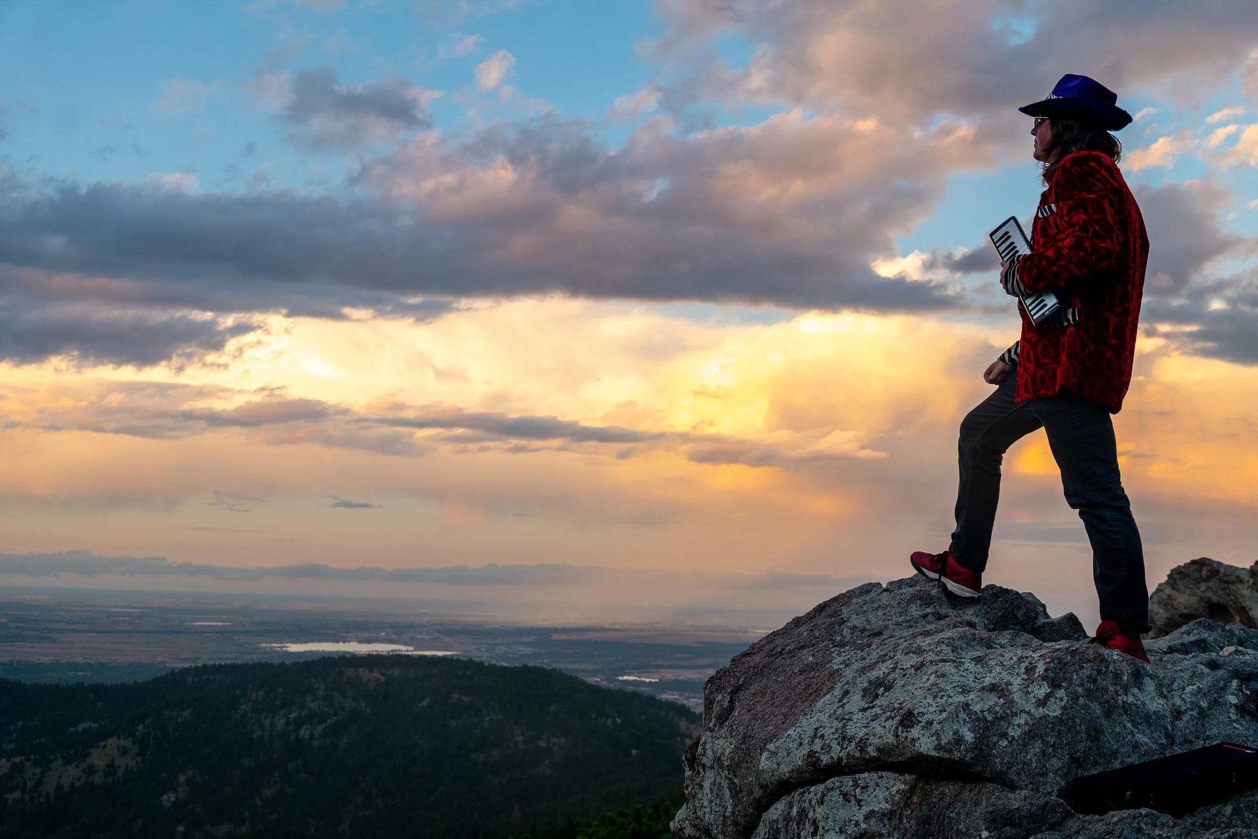 Person standing on a large rock on a mountain with a landscape and sunset sky in the background.