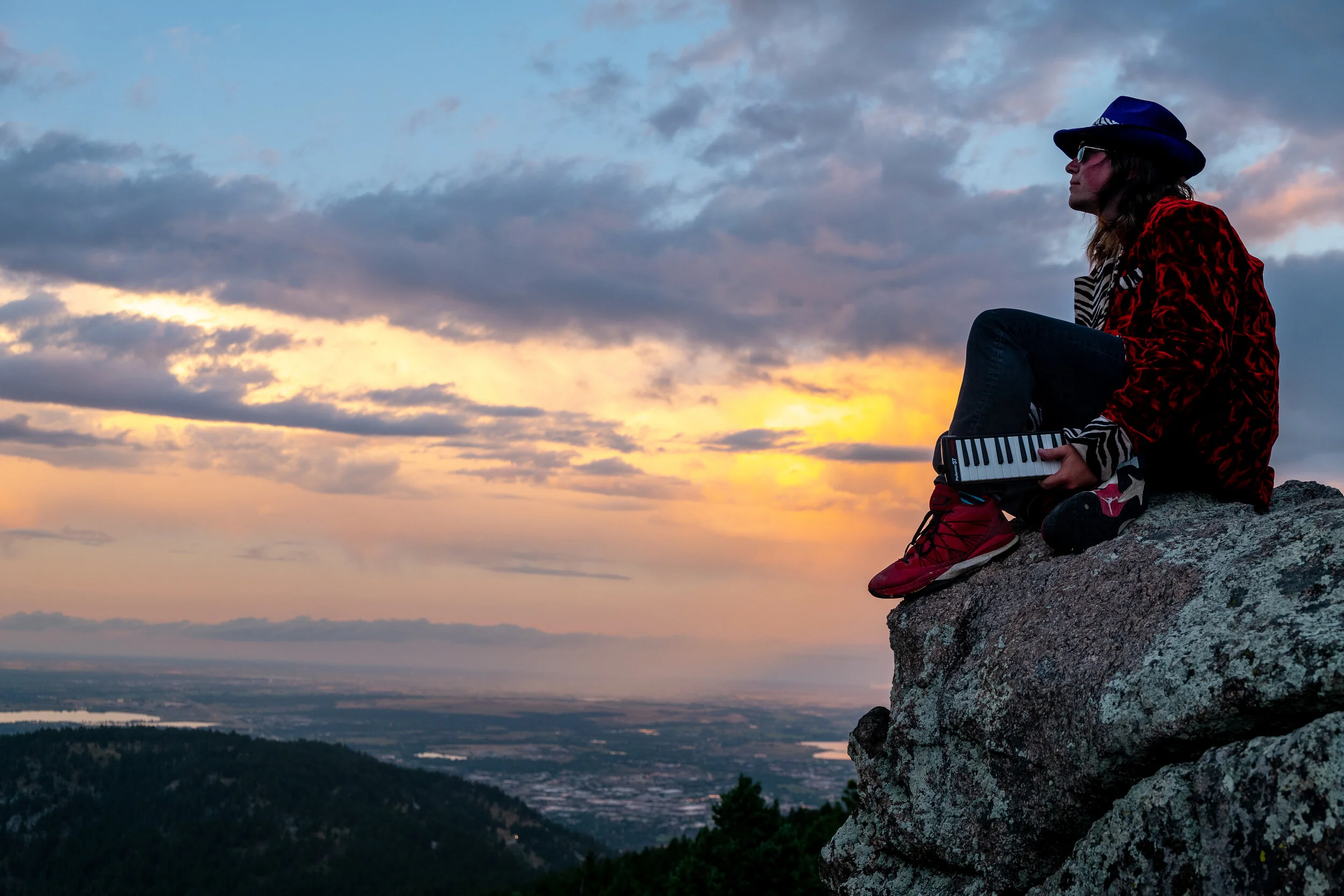A person sitting on a rock ledge overlooking a sunset landscape, holding a small keyboard, wearing a blue hat, sunglasses, a red patterned jacket, and sneakers.