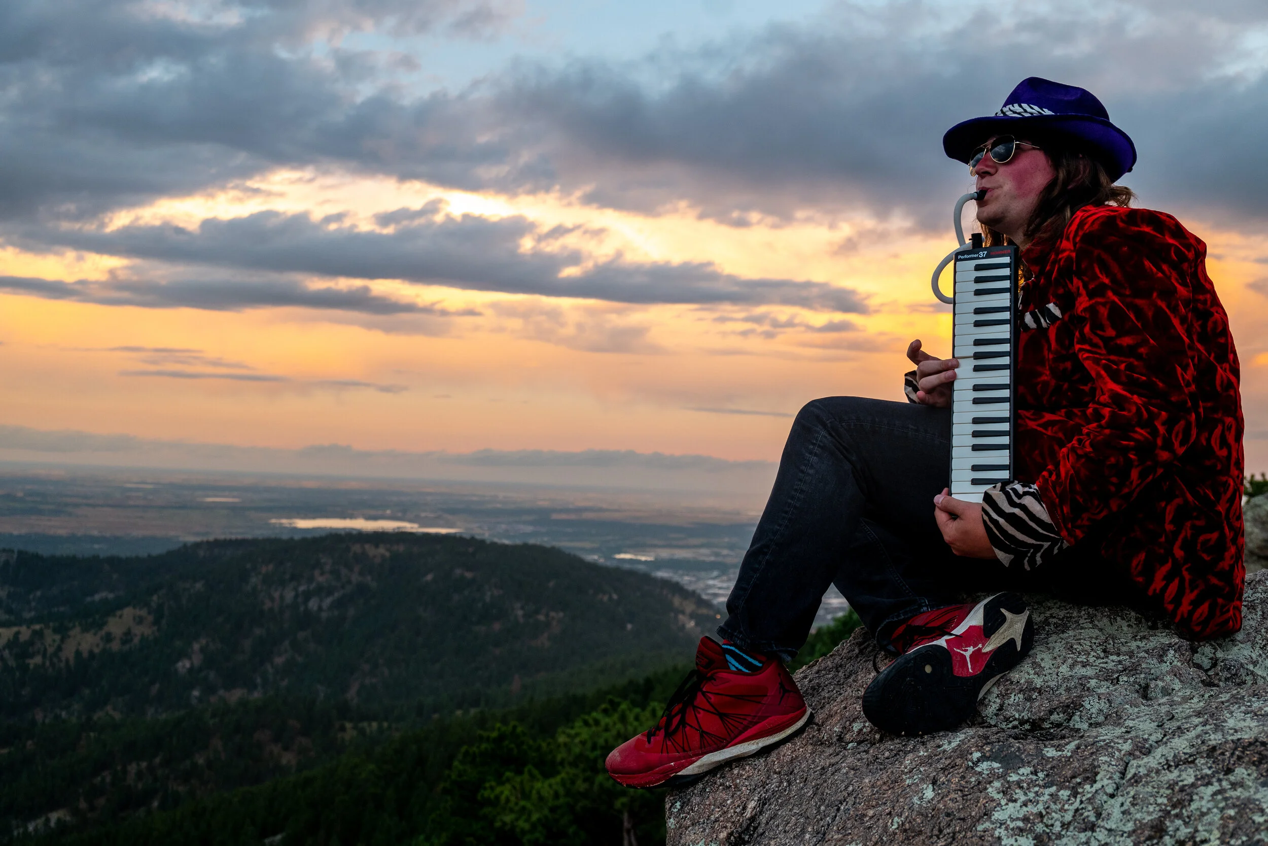Person sitting on a rock playing a melodica, wearing a blue hat, sunglasses, red velvet jacket, and red sneakers, with a sunset and landscape in the background.